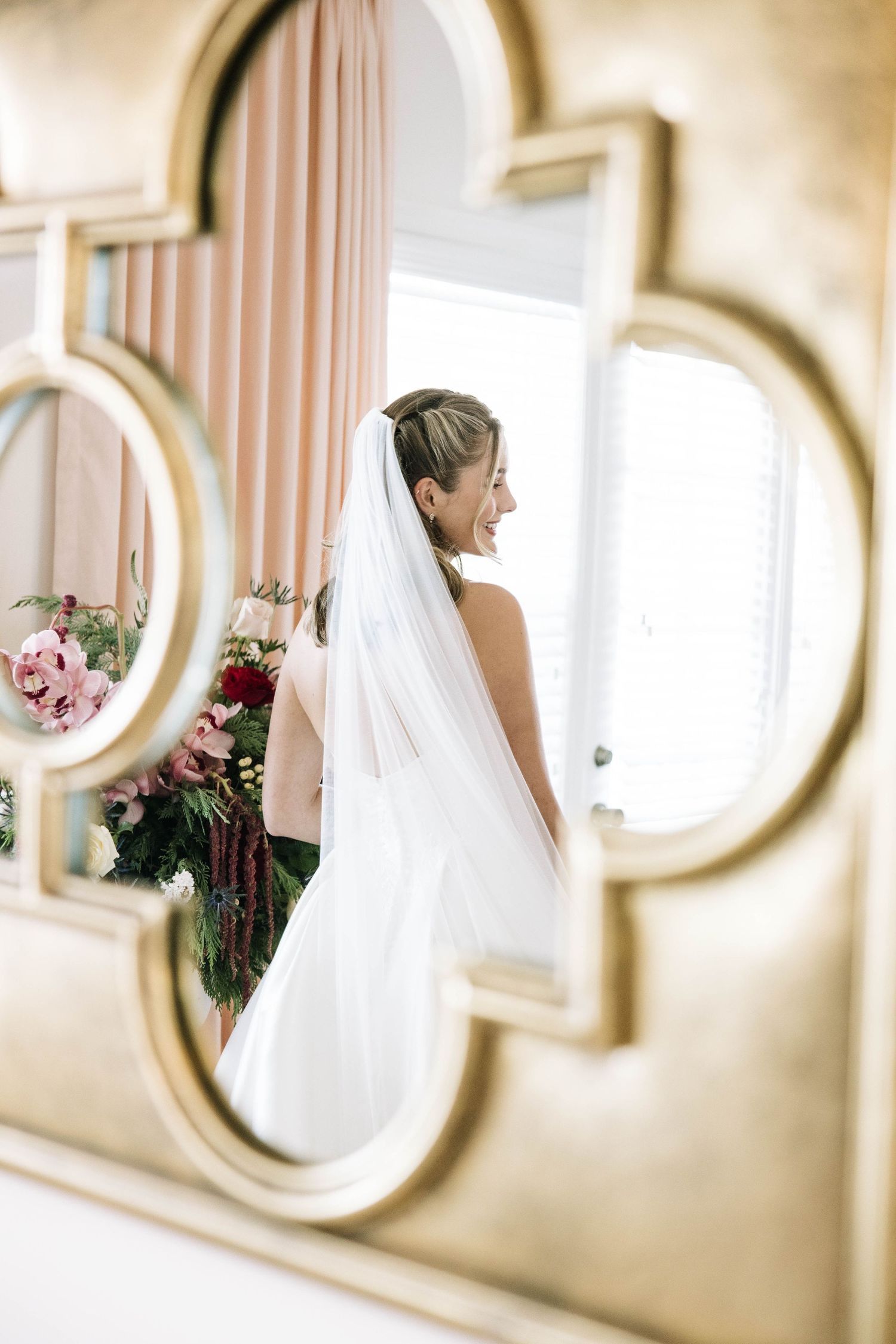 Bride in white veil smiles holding bouquet, reflected in ornate gold mirror.