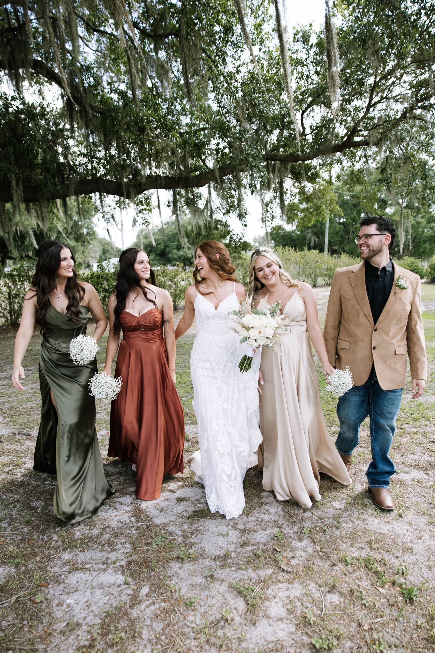 Bride in lace gown walks with bridesmaids in earth-tone dresses under Spanish moss-draped trees.
