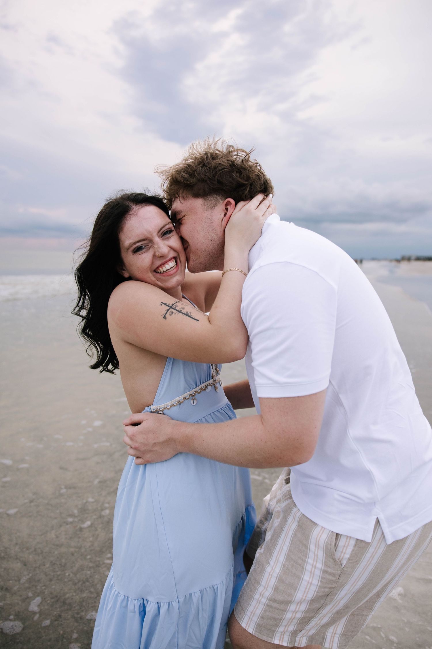 Joyful couple embracing on a beach, woman in blue dress laughing as man kisses her cheek by the ocean.