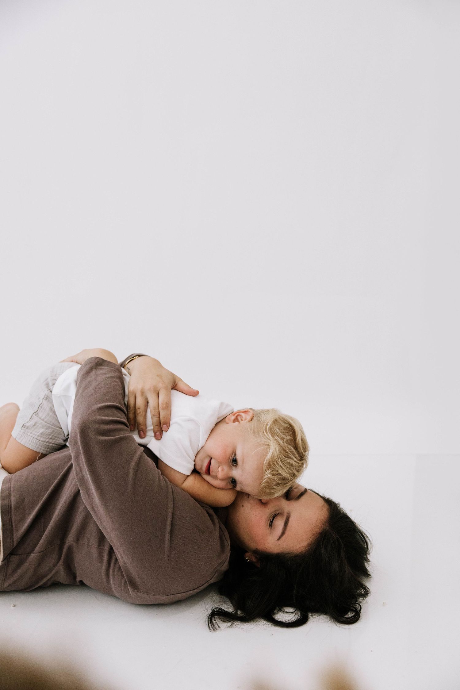 Mother lying on white background cuddling and embracing her laughing blonde toddler child with joy.