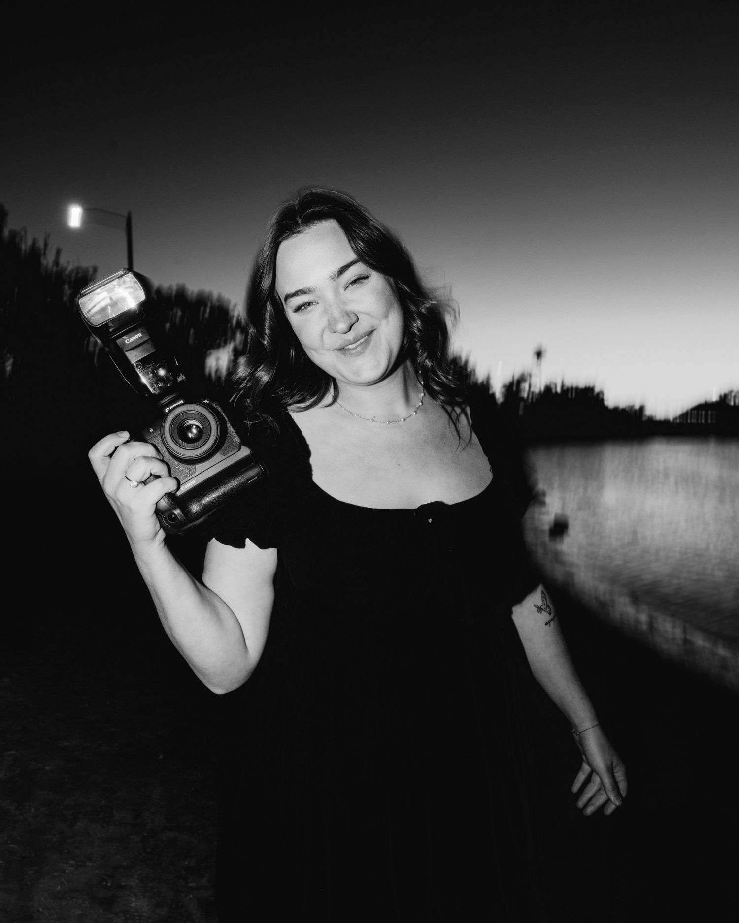 Black and white photo of smiling female photographer holding flash camera at dusk.