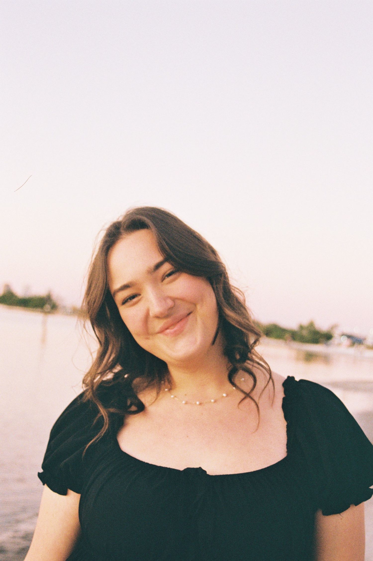 Young woman with long brown hair smiling warmly at sunset near a waterfront wearing a black dress.