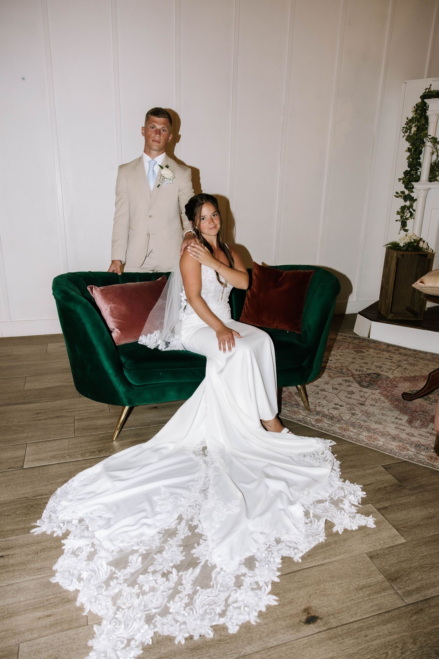 Bride in lace gown with long train sits on green velvet sofa as groom stands behind her indoors.