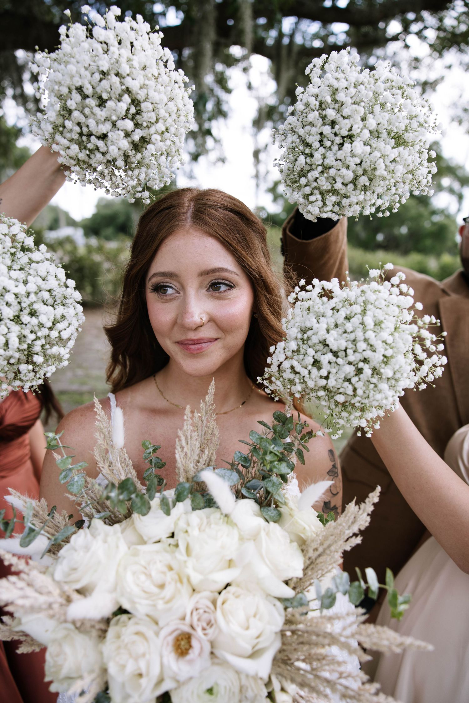 Bride surrounded by bridesmaids holding baby's breath bouquets, smiling softly while holding a white rose bridal bouquet outdoors.
