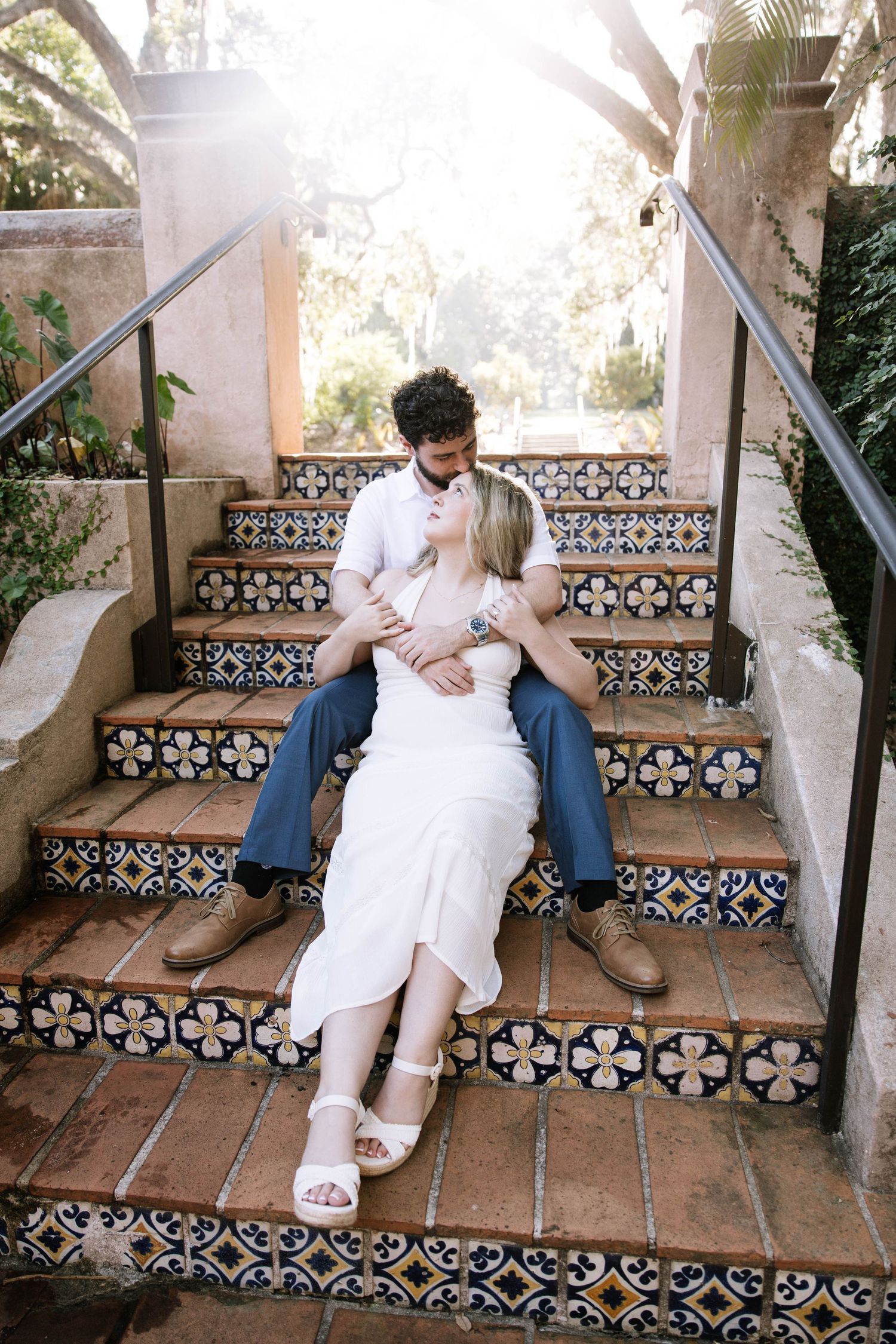 Couple sitting on decorative tiled steps embracing in sunlit garden setting.