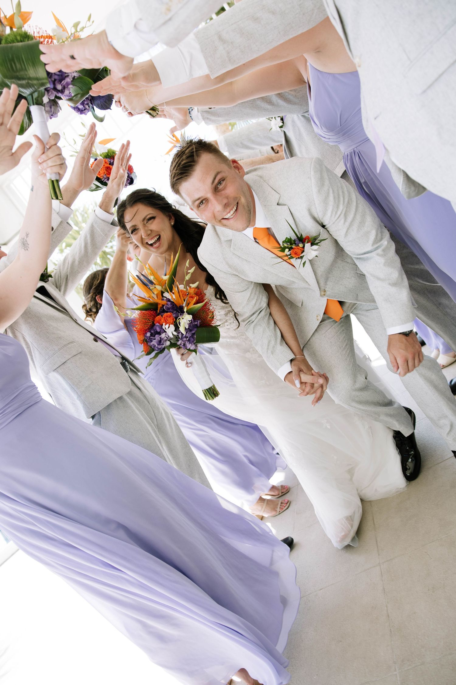 Joyful bride and groom walk through cheering wedding party in lavender dresses, bride holding tropical bouquet.