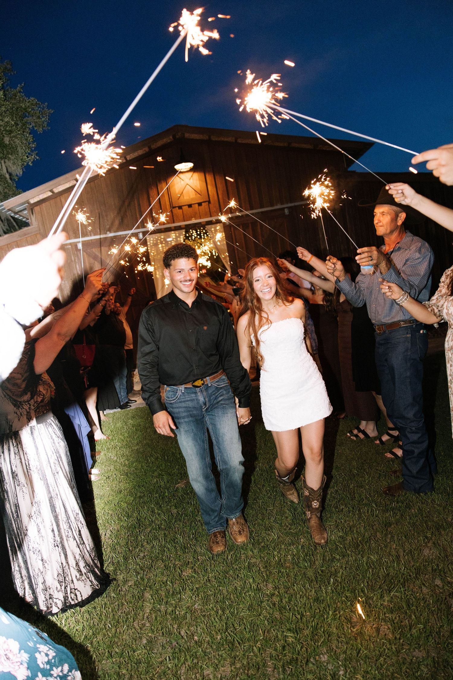 Newlyweds walk through sparkler tunnel at outdoor evening wedding reception with guests celebrating.
