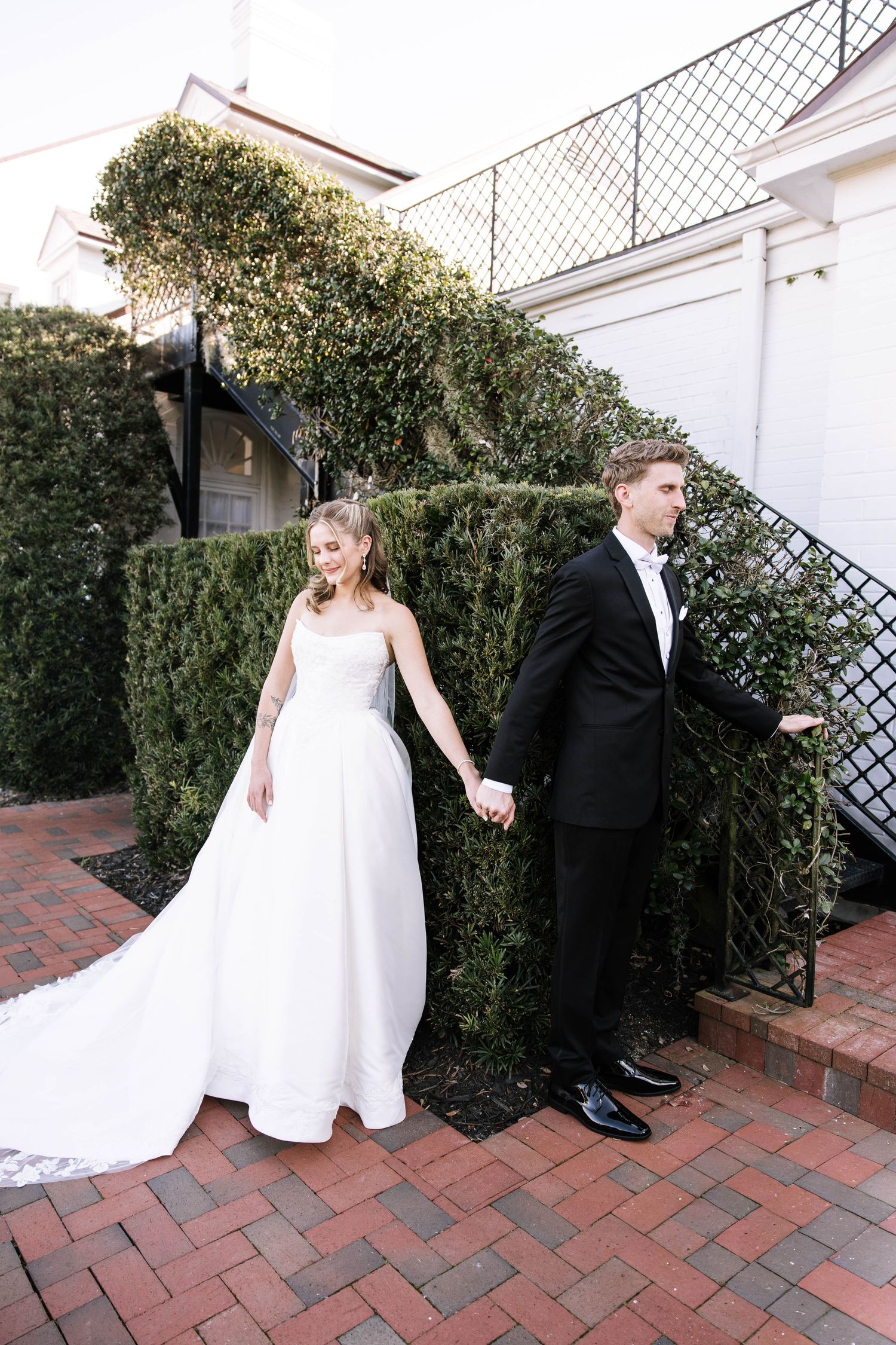 Bride and groom hold hands facing opposite directions beside ivy-covered wall on brick courtyard.