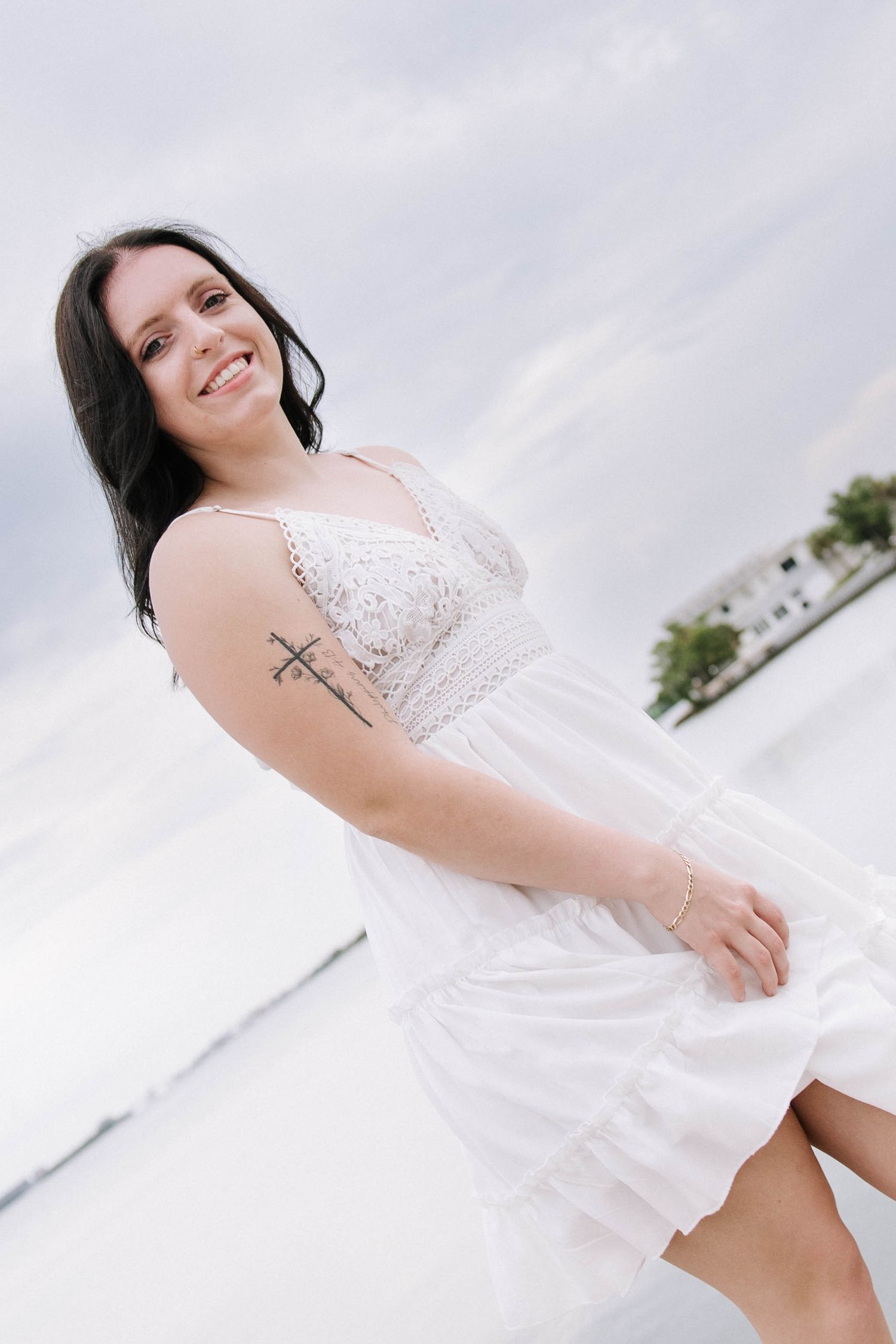Smiling brunette woman in white sundress with arm tattoo posing outdoors on bright overcast day near water.Photographers at Anna Maria Islan