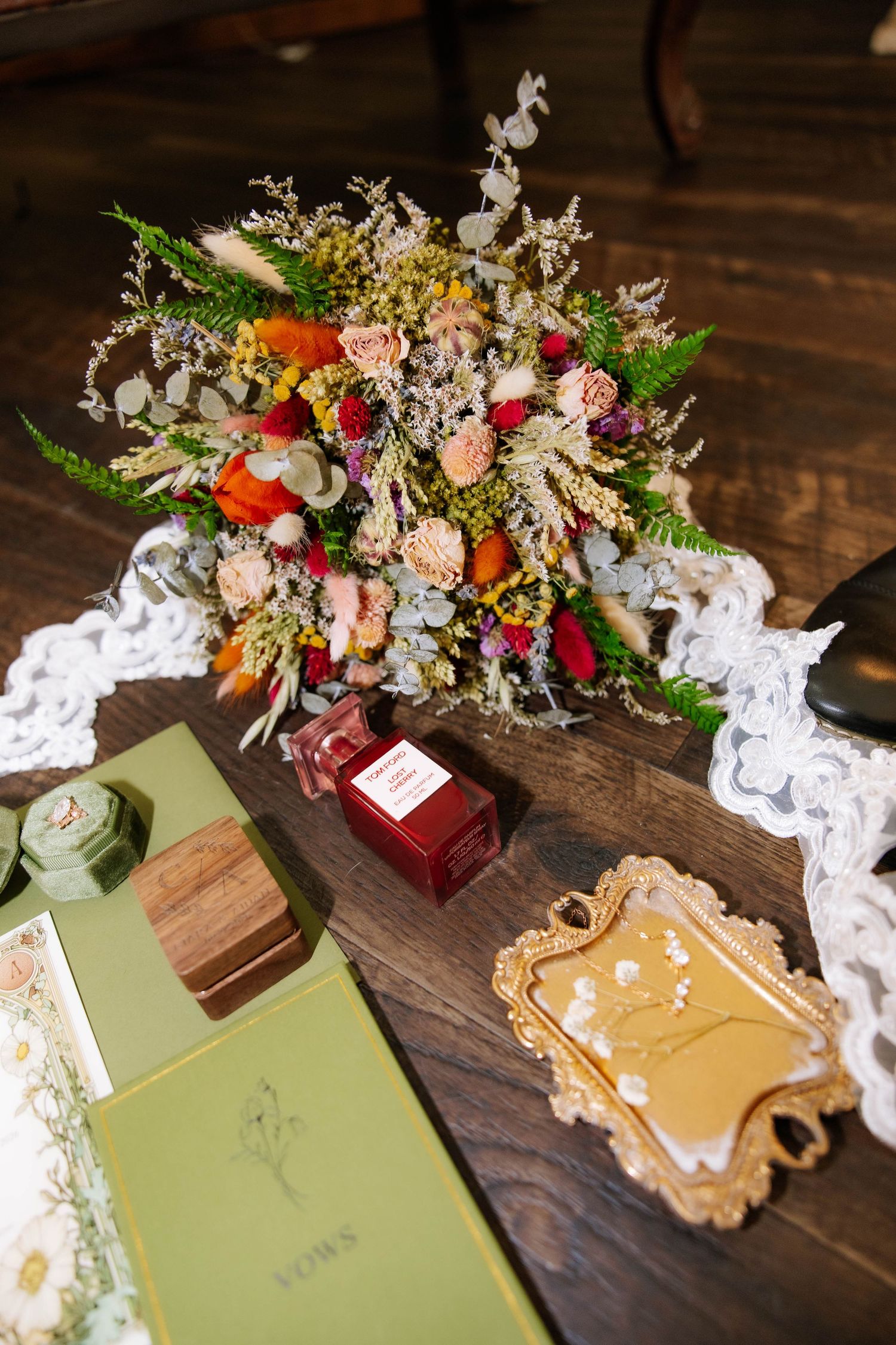 A rustic wedding flat lay featuring a colorful floral bouquet, perfume bottle, vow books, and gold tray on a wooden table.