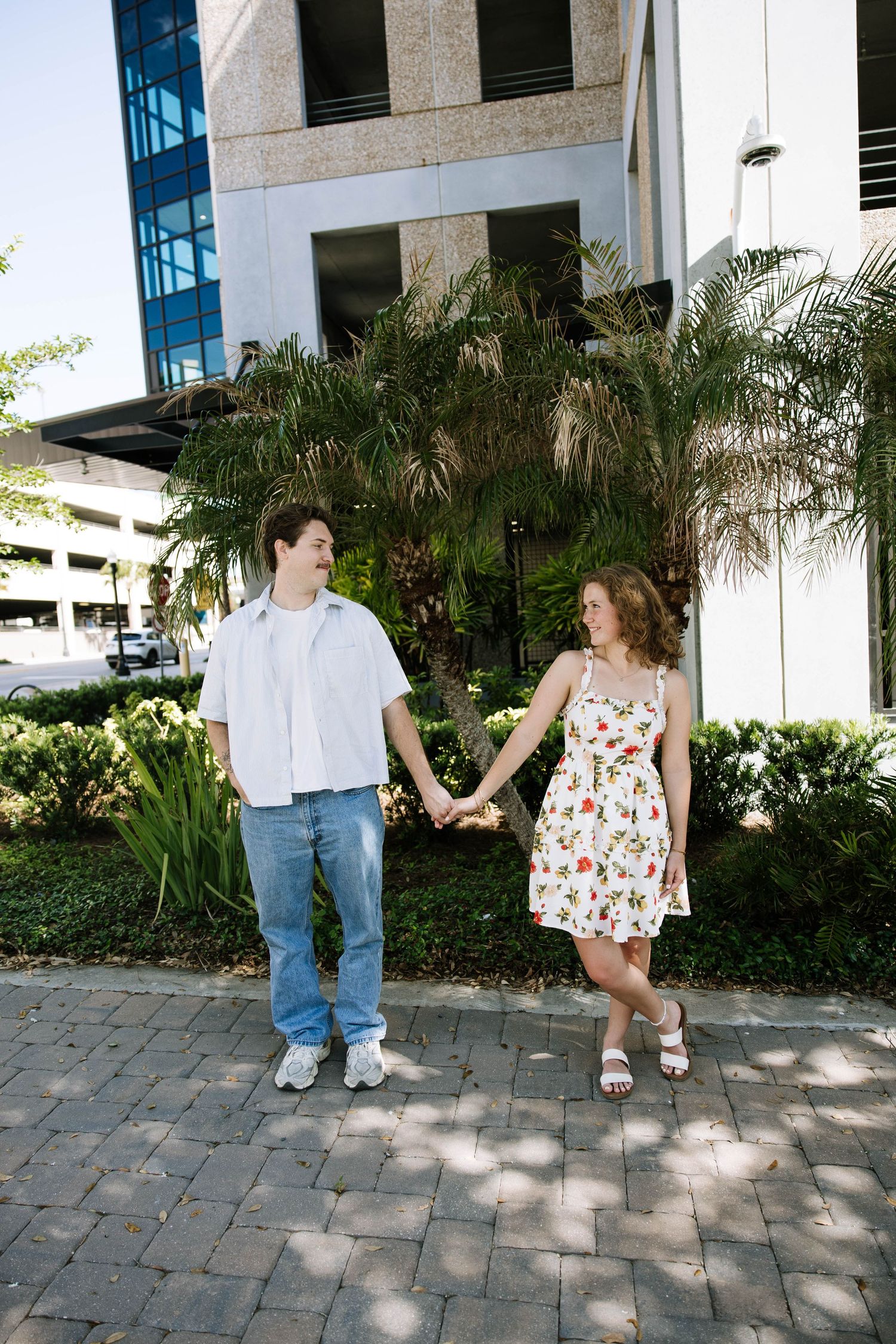 Young couple holding hands and smiling at each other while walking on a brick path outside a modern building.
