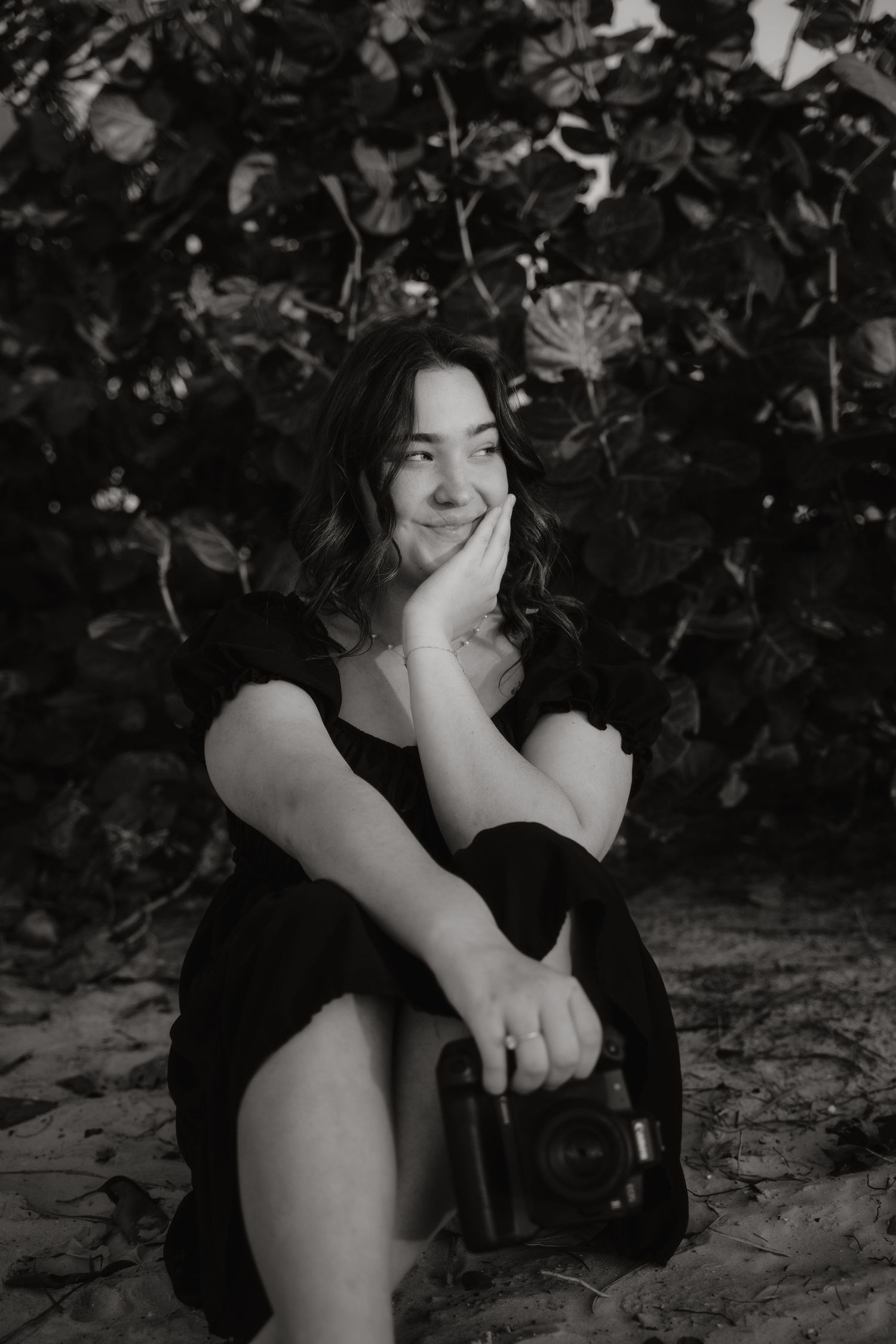 Black and white portrait of a young woman smiling, holding a camera outdoors.