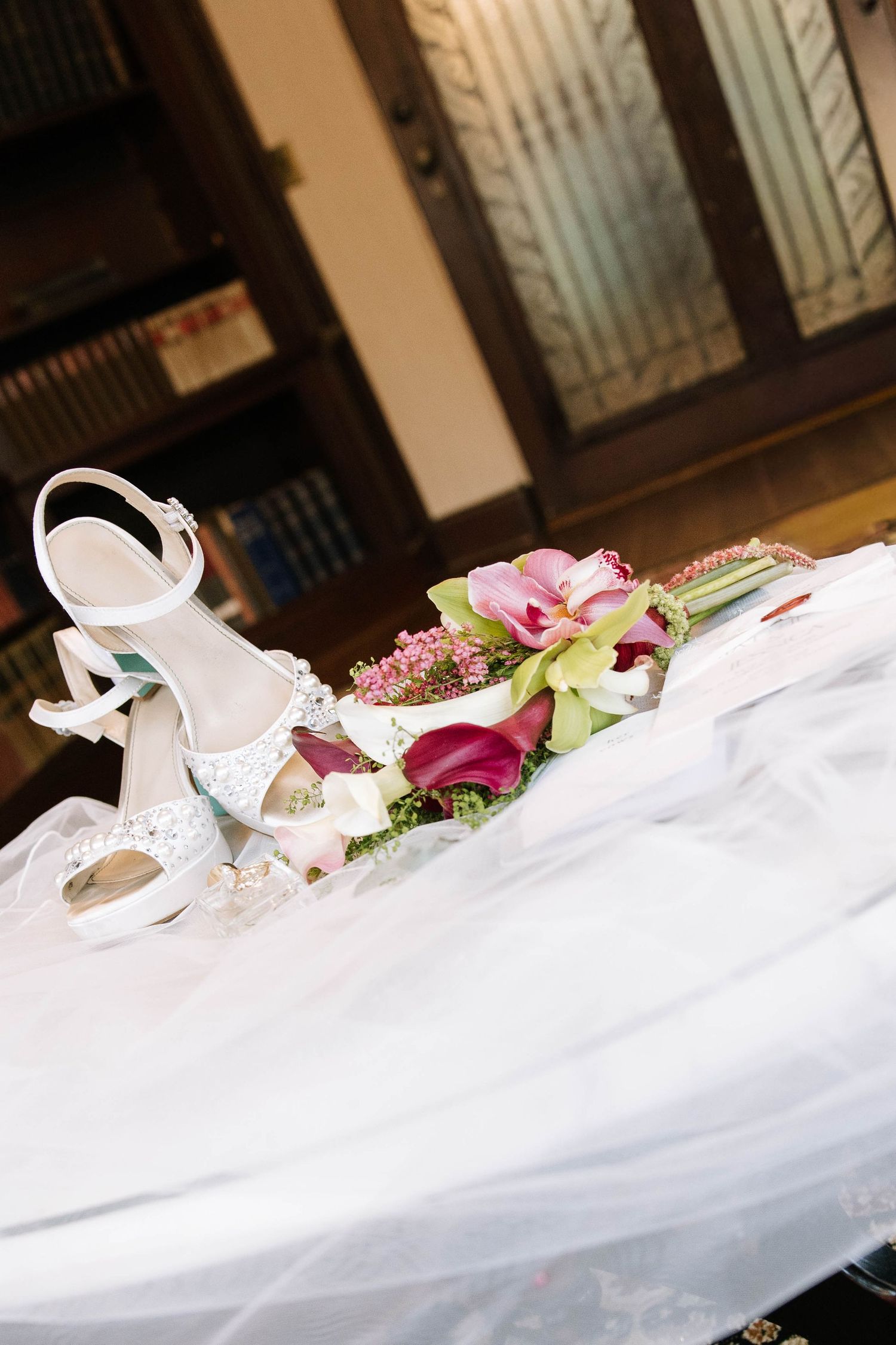 White bridal heels and pink calla lily bouquet resting on a wedding veil on a table.