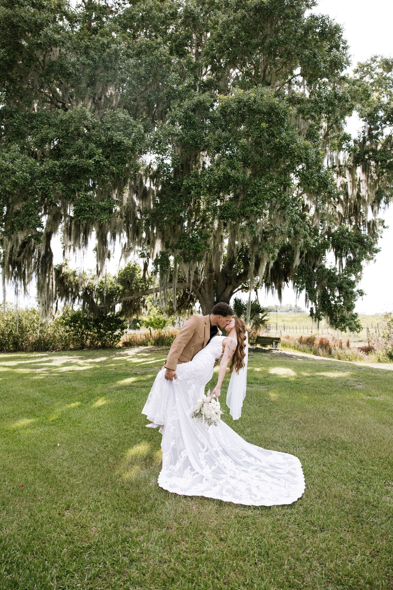 A groom dips his bride in a white lace dress for a romantic kiss beneath a large moss-draped oak tree.