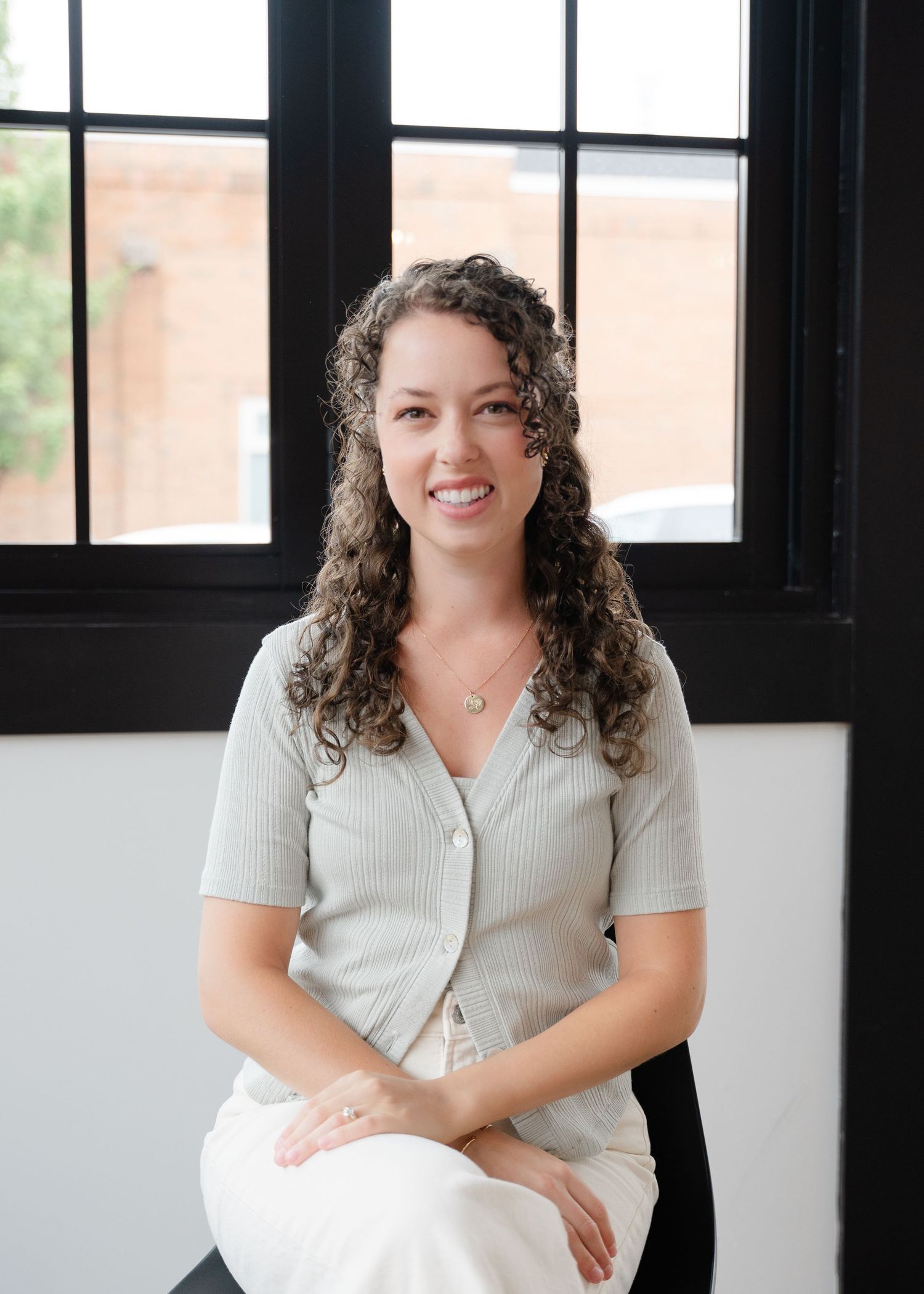 Smiling woman with curly hair in sage green top seated by large black-framed windows.