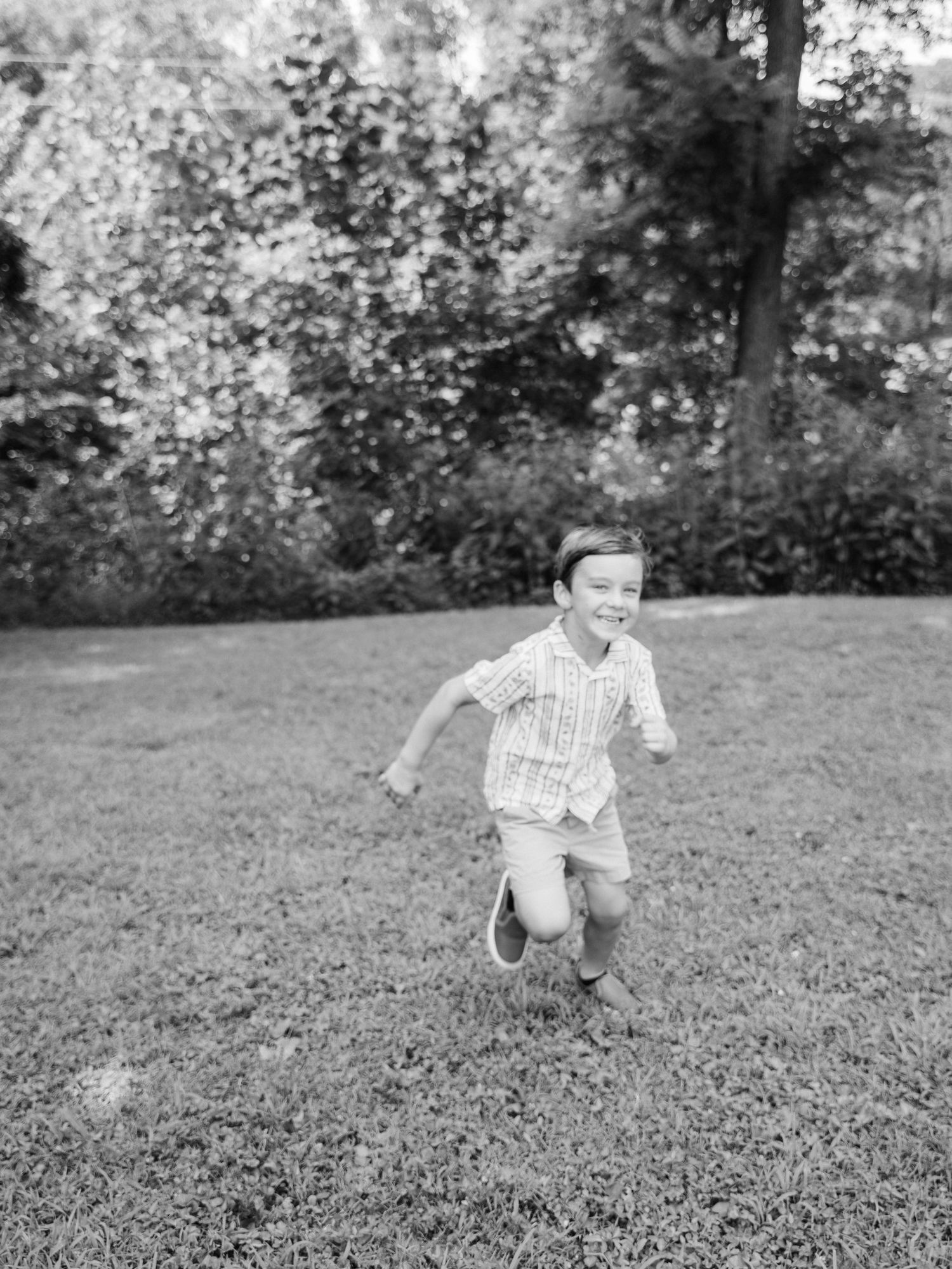 A joyful young boy running and smiling across a lush green lawn with trees in the background.