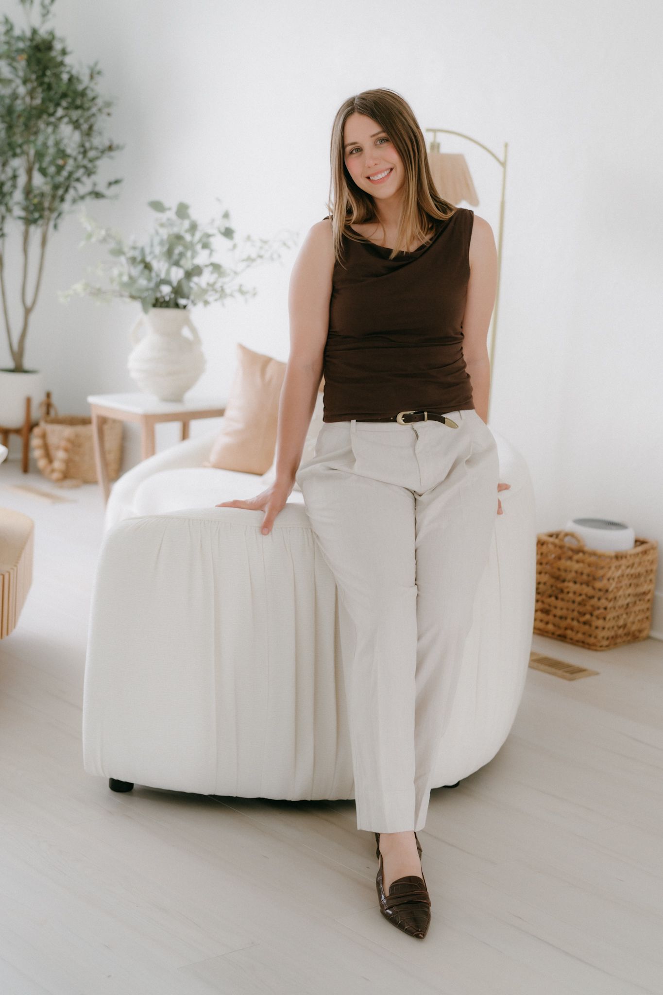 Smiling brunette woman in brown top and white pants posing beside a modern white chair.