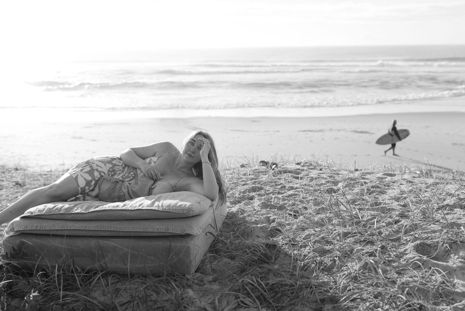 Mother breastfeeding her baby on the beach as a surfer passes in the background