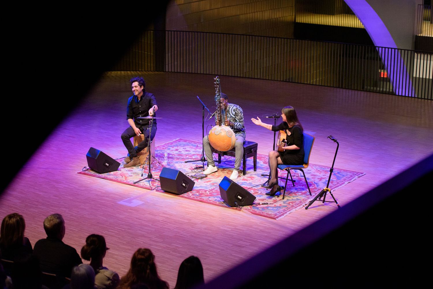 Two musicians perform on a purple-lit stage with an oud instrument, seated on a decorative rug in an intimate concert hall.