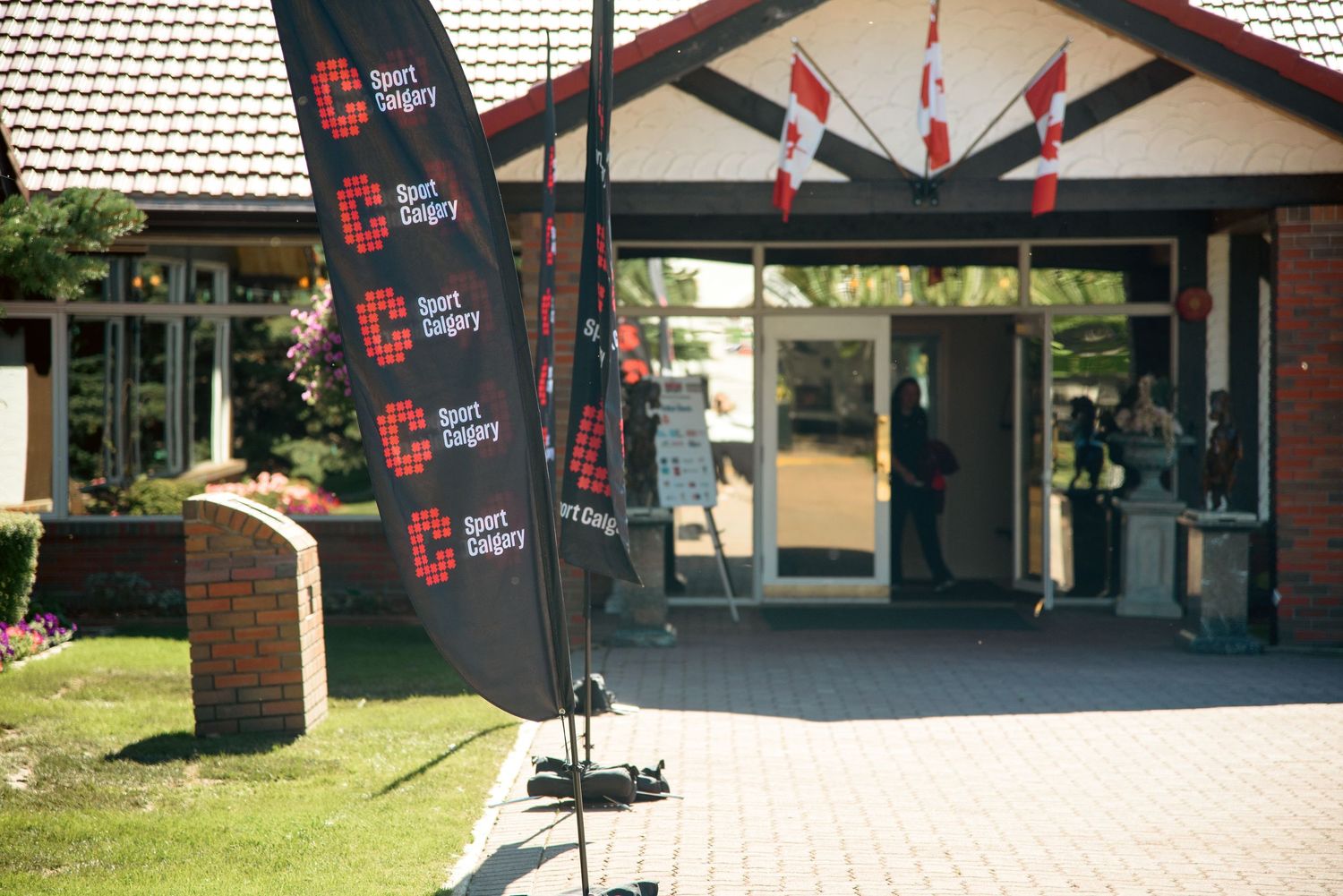 Sport Calgary branded feather flags displayed outside a brick community recreation center entrance on a sunny day.