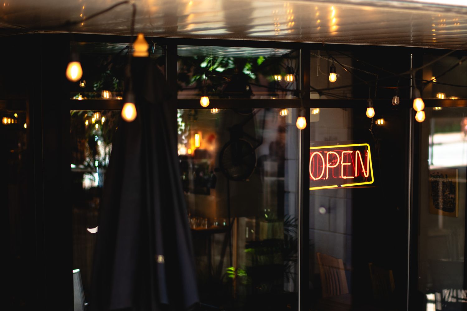 Neon open sign glowing in dark restaurant window with warm Edison bulb lights inside.