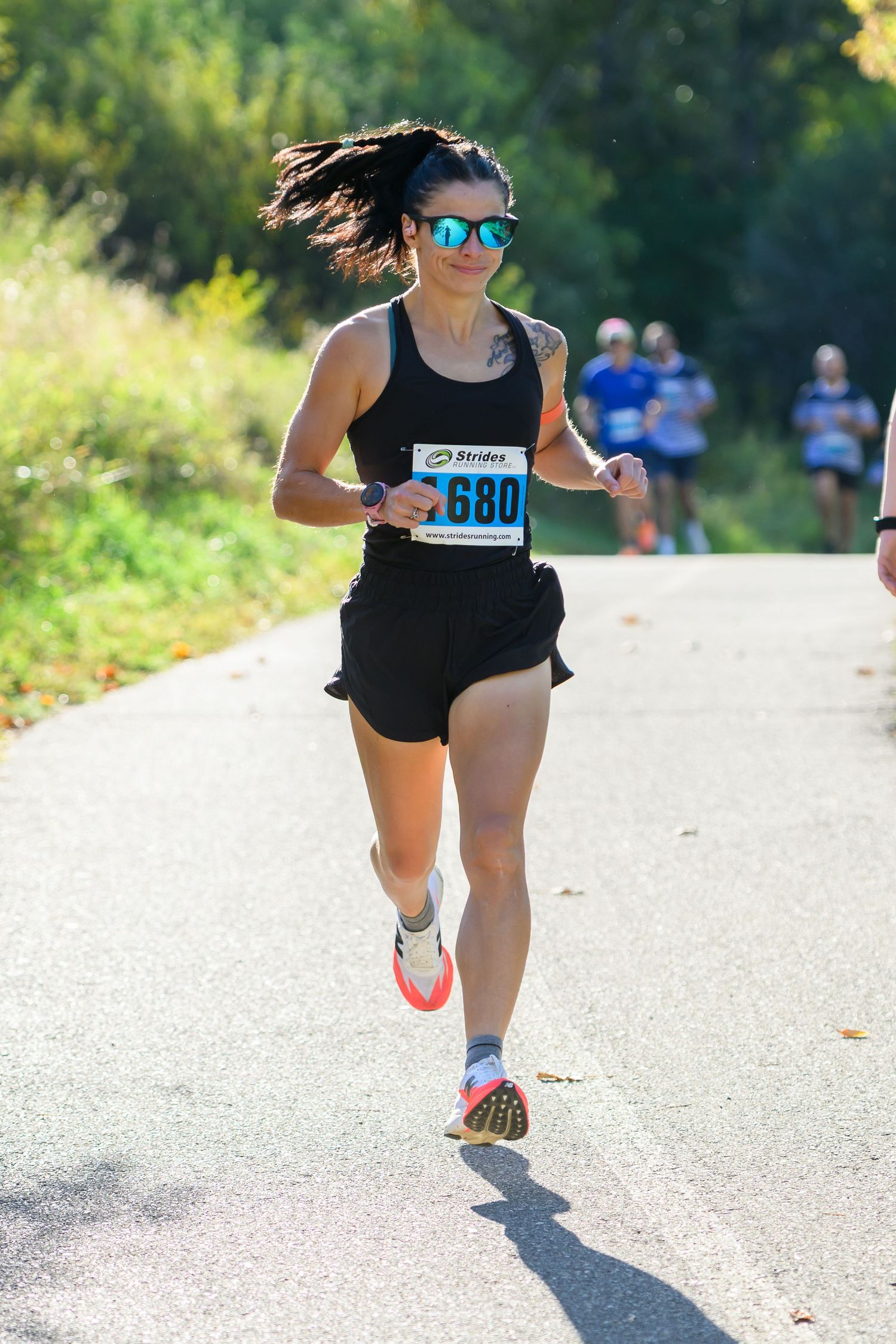 Woman in black tank top and shorts, wearing sunglasses, running a road race with bib number 680 on a sunny day.