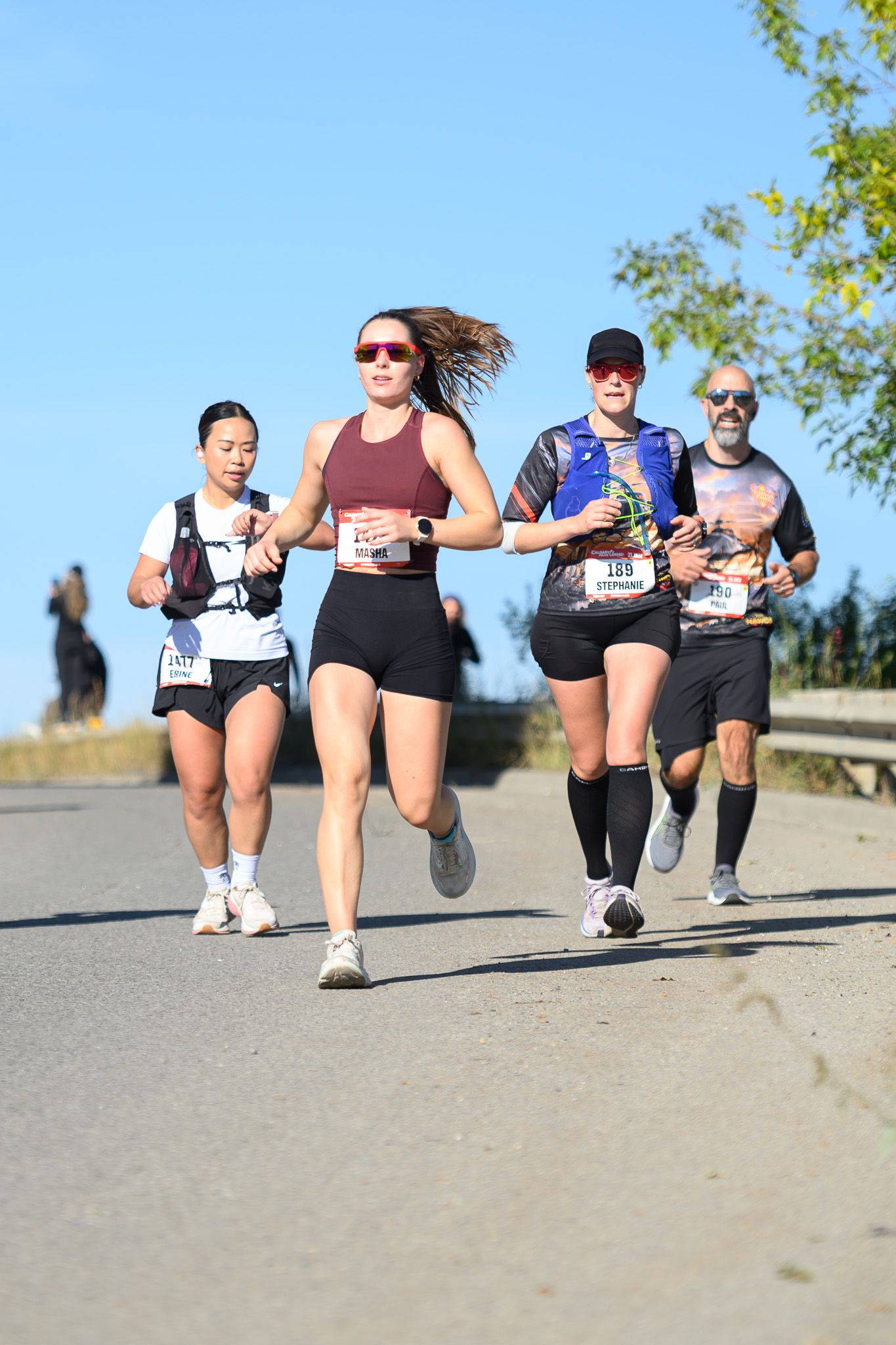 Four runners competing in an outdoor road race on a sunny day, wearing race bibs and athletic gear.