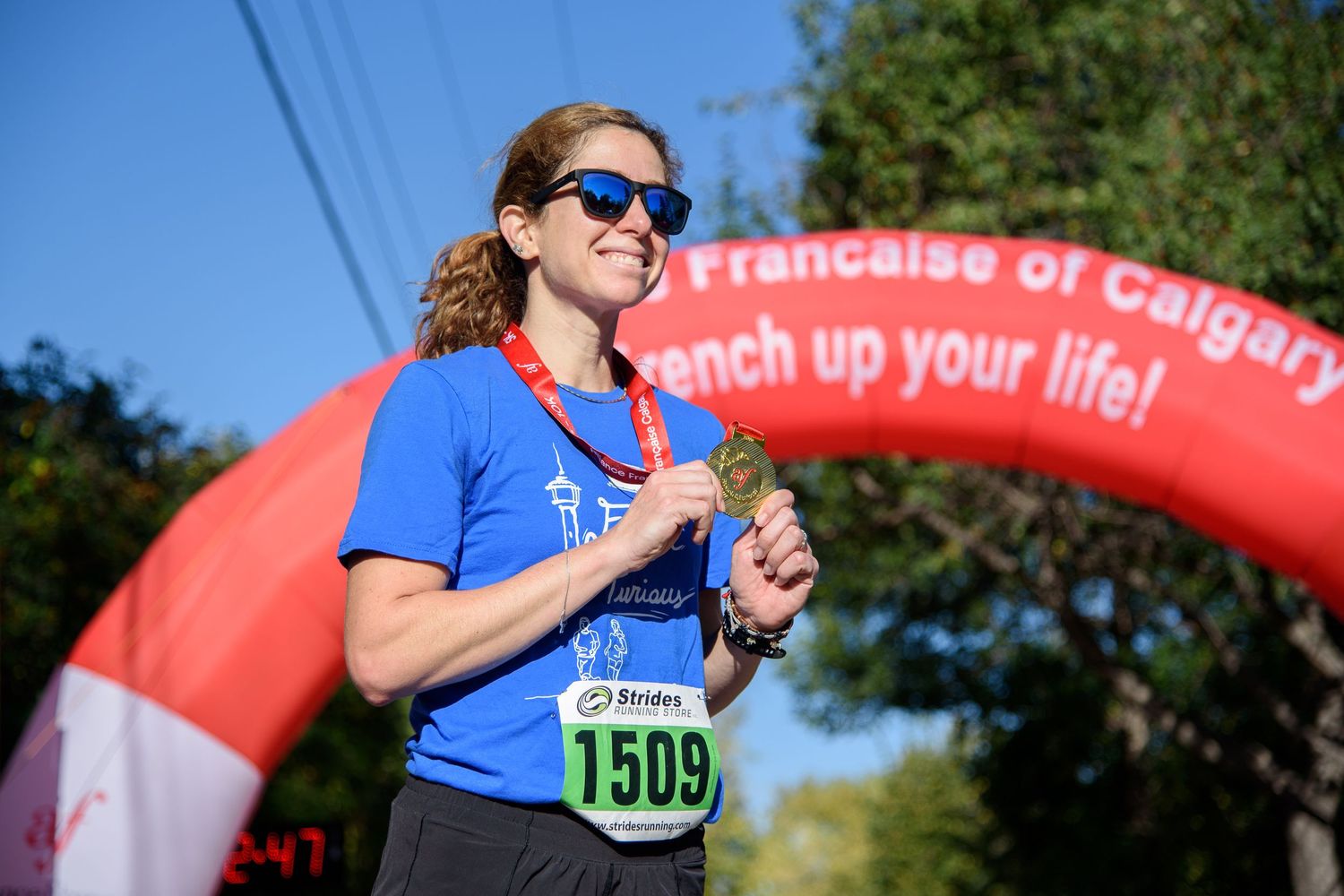 Woman runner holding medal at finish line of Calgary French race wearing bib number 1509 on sunny day.