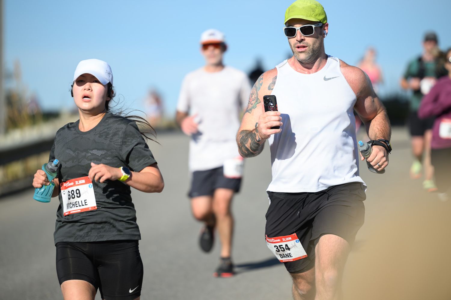 Runners competing in a road race, wearing numbered bibs, on a sunny day with clear skies in the background.