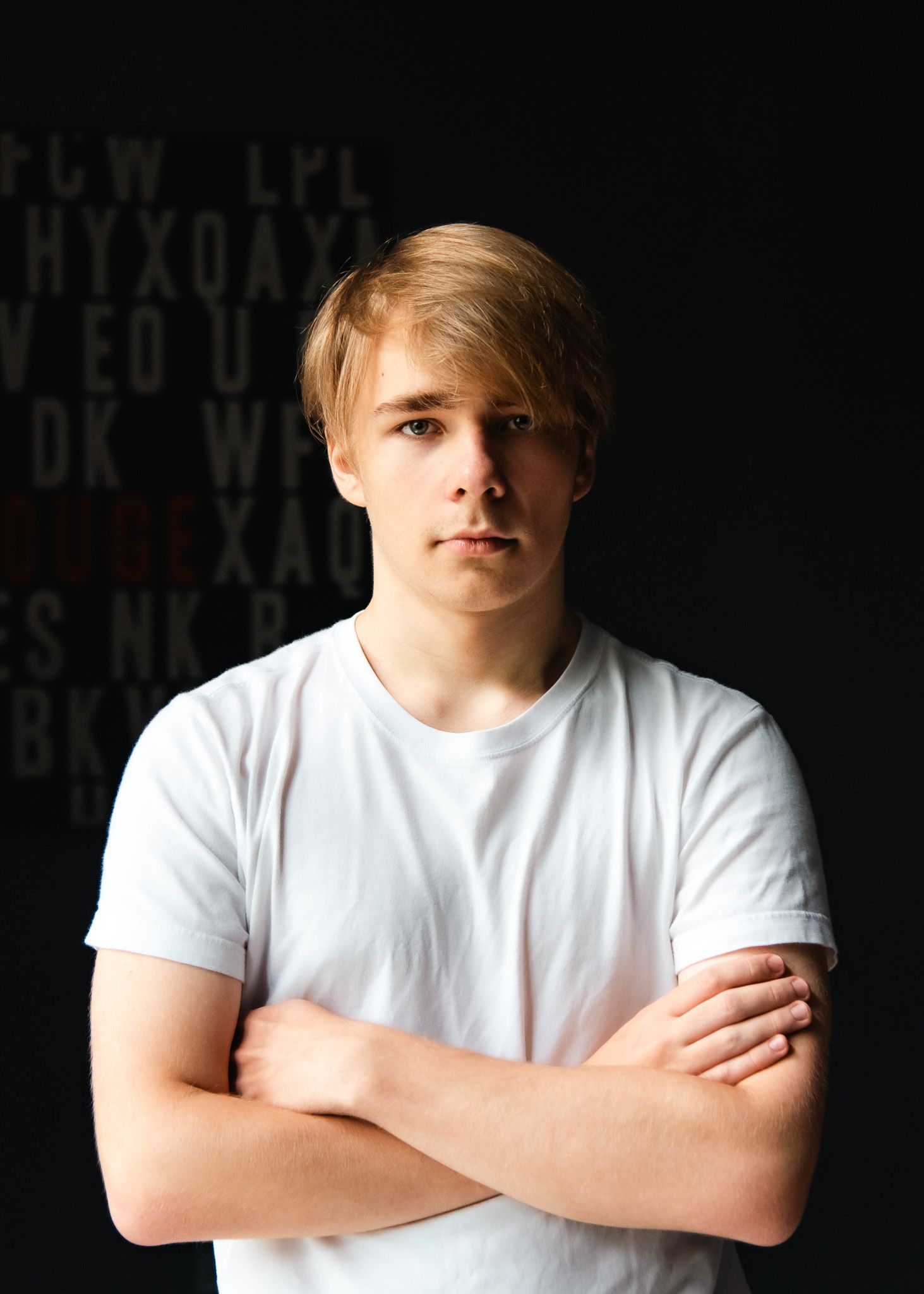 Young blonde man in a white t-shirt stands with arms crossed against a dark background.