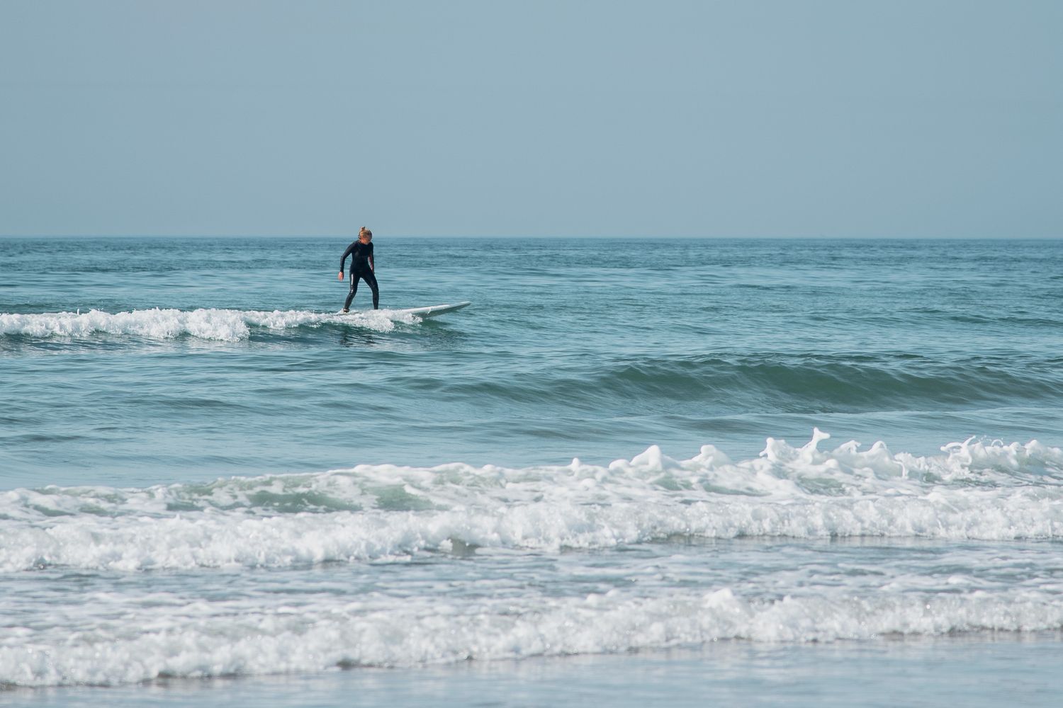 Surfer riding small ocean waves on a clear day at a sandy beach.