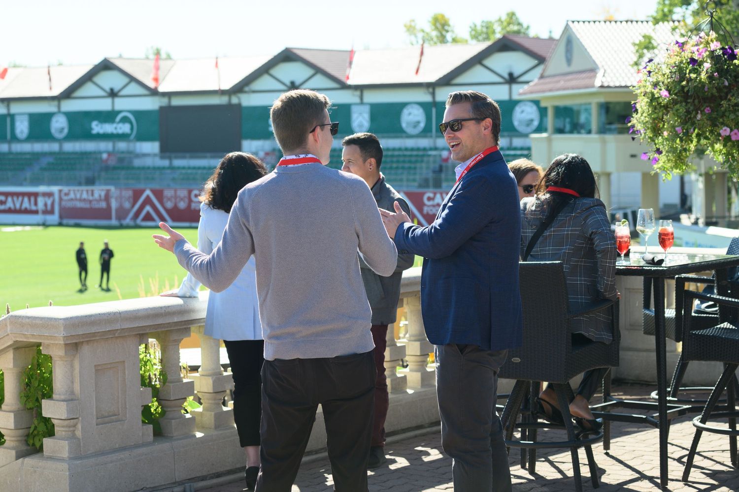 Group of people networking outdoors near a sports field on a sunny day, with green stadium visible in background.