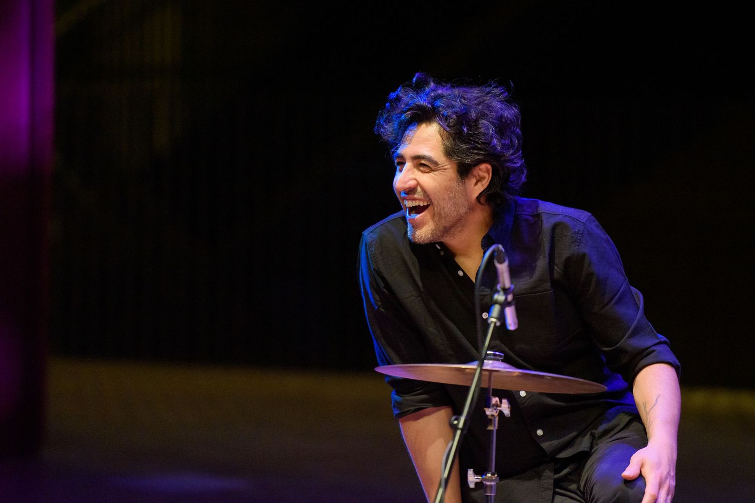 Smiling male musician performing on stage near a microphone and cymbal under warm stage lighting.