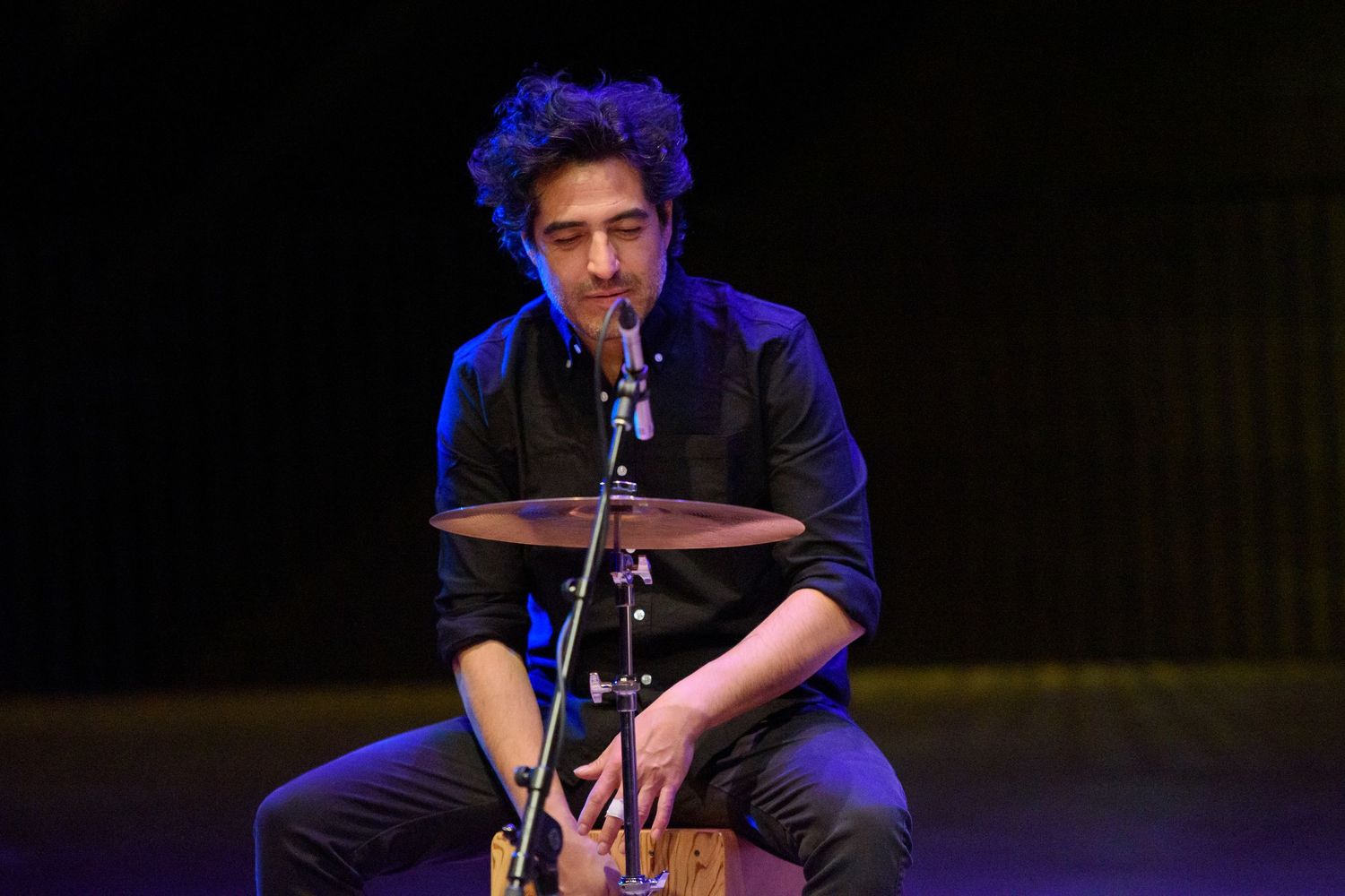 Male musician playing cajon drum on stage with cymbal and microphone stand in a live performance setting.