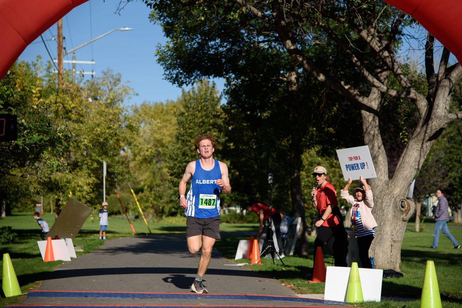 Runner crossing finish line at outdoor 5K race event, wearing blue singlet with race bib, trees in background.