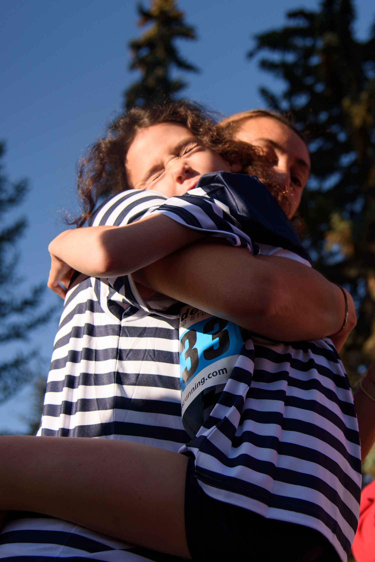 Two people in matching navy striped shirts share a warm embrace outdoors with pine trees and blue sky behind them.