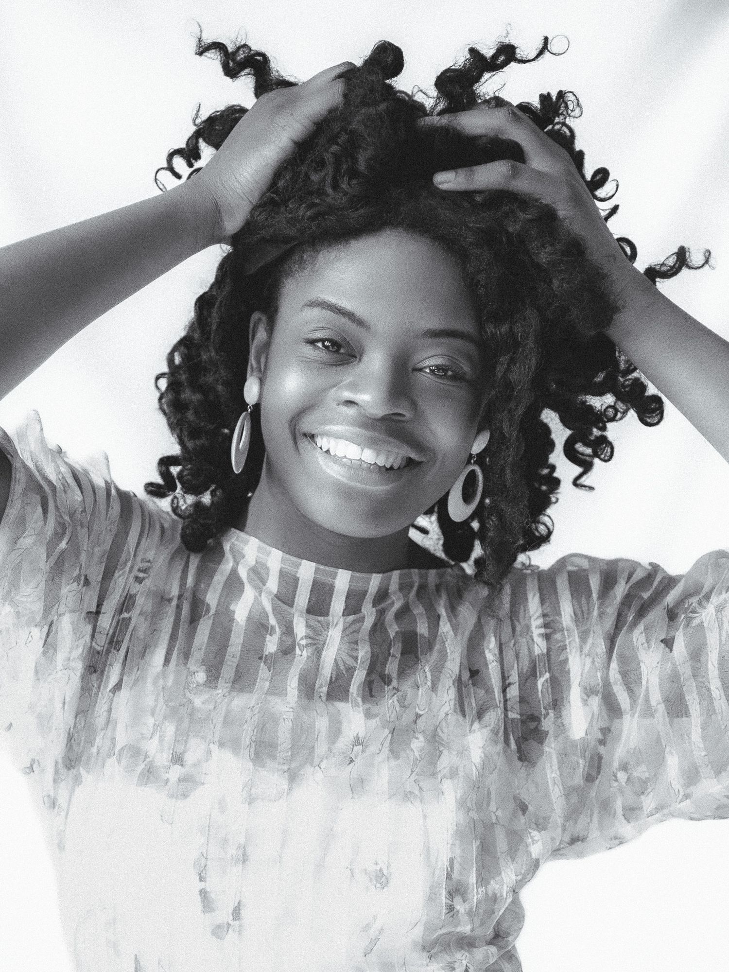 Black and white portrait of smiling young Black woman with curly hair holding head with both hands.