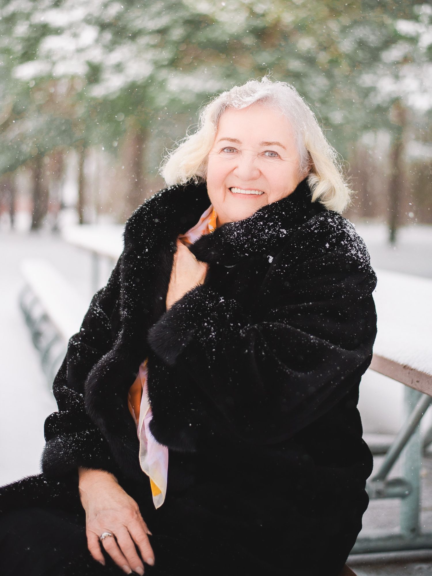 Smiling older woman with white hair in black fur coat sitting on bench in snowy winter park.