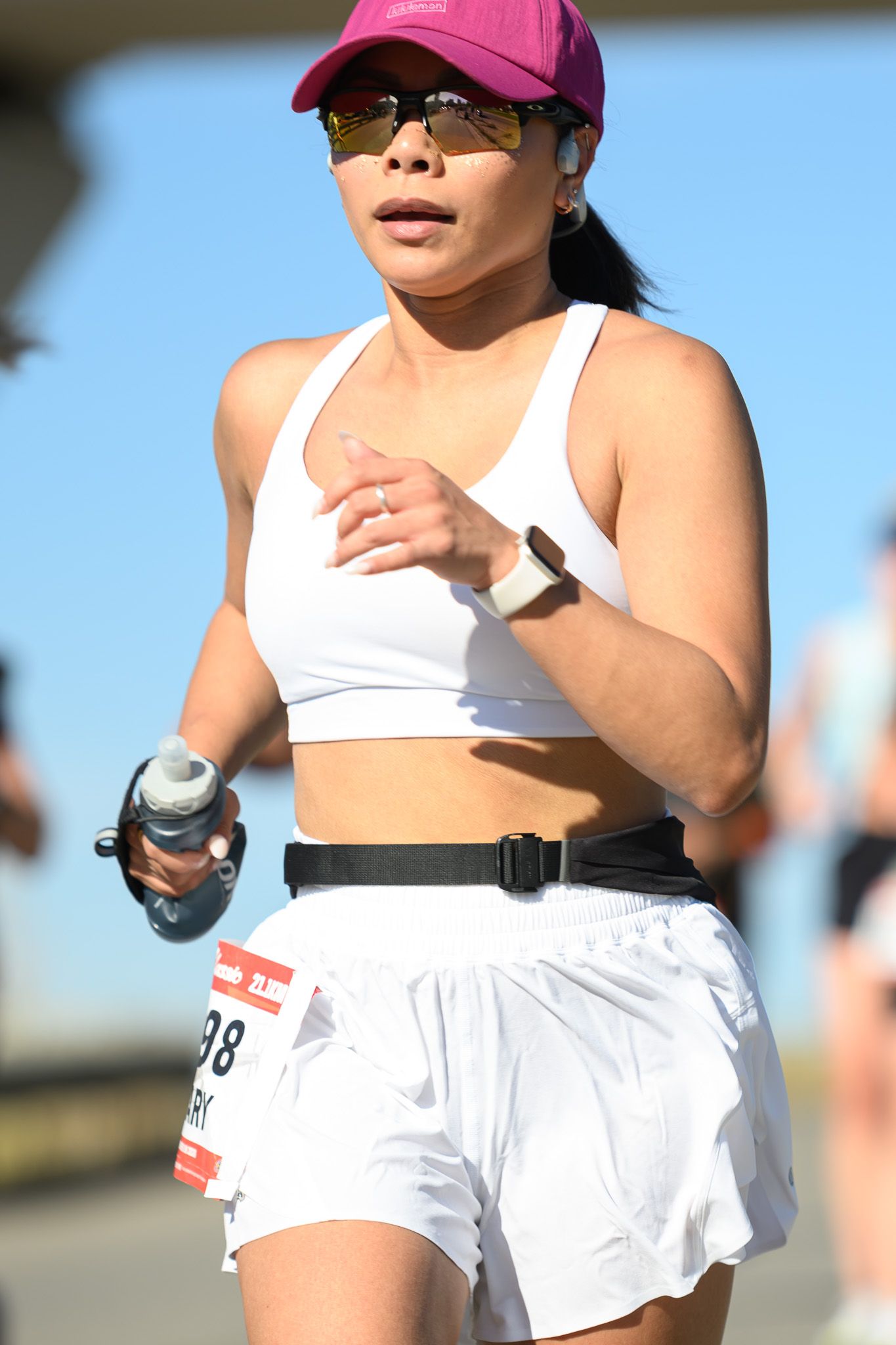 Woman in white sports bra and shorts with race bib running a marathon wearing pink cap and sunglasses.