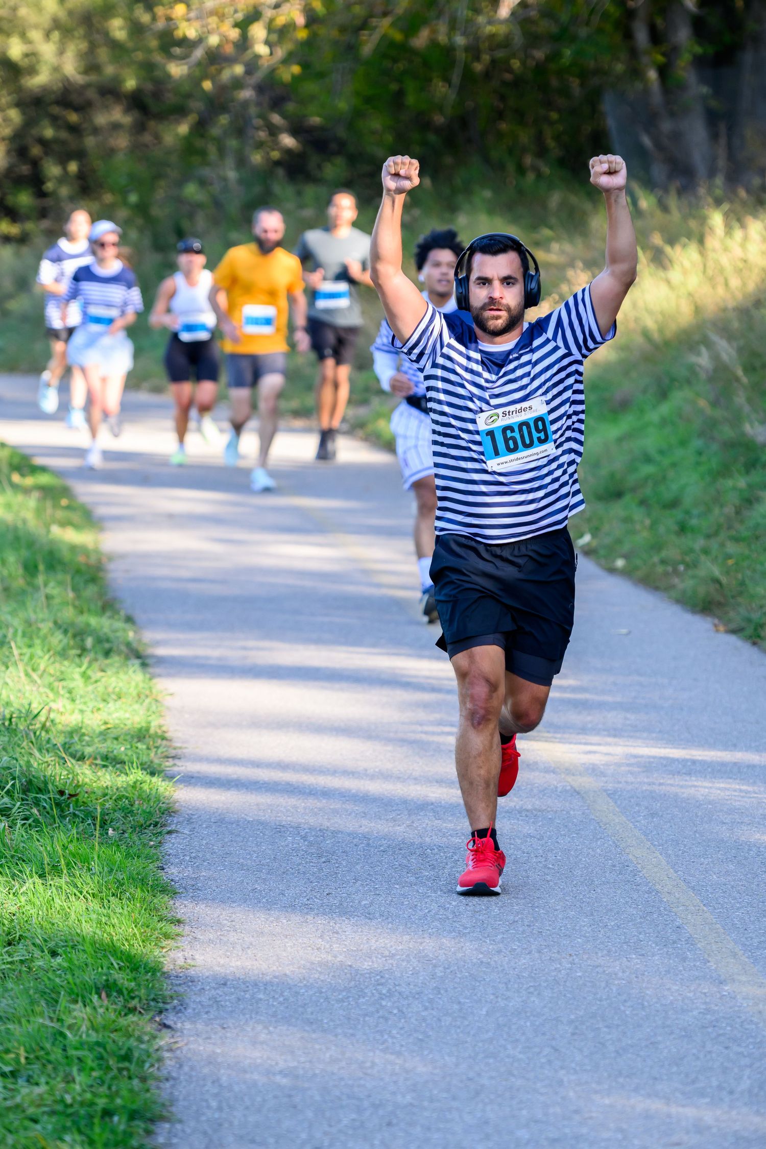 Male marathon runner #1609 raises fists in triumph wearing striped shirt and red shoes on tree-lined race course.