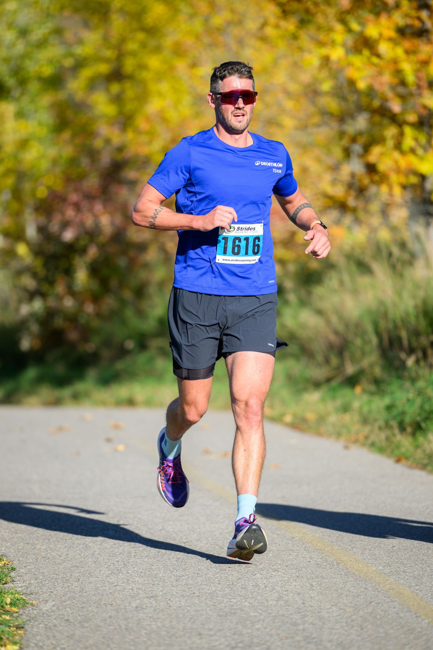 Male runner wearing bib 1616 races along a paved path surrounded by autumn foliage.