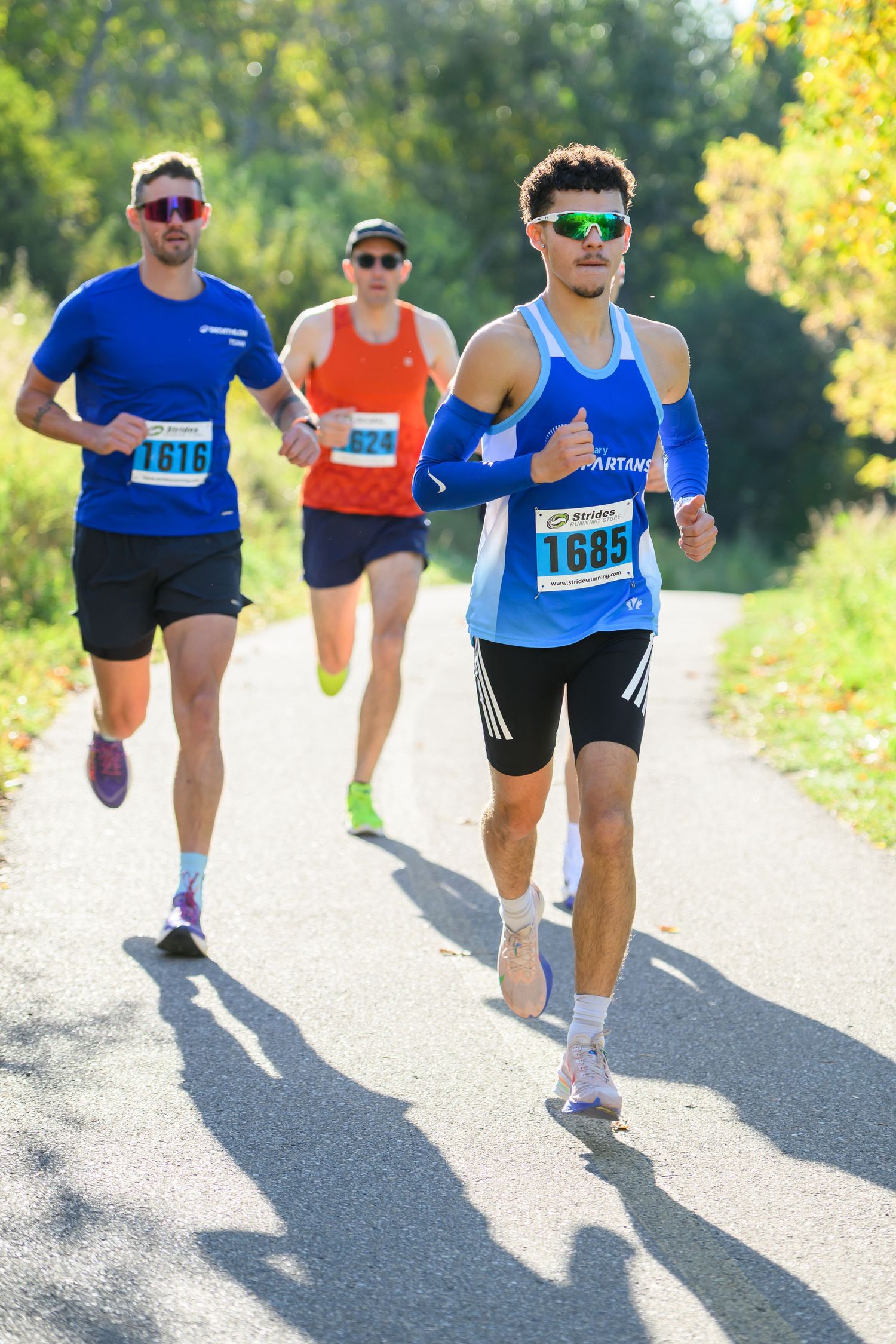 Three male runners wearing numbered bibs compete in an outdoor road race through a tree-lined path on a sunny day.