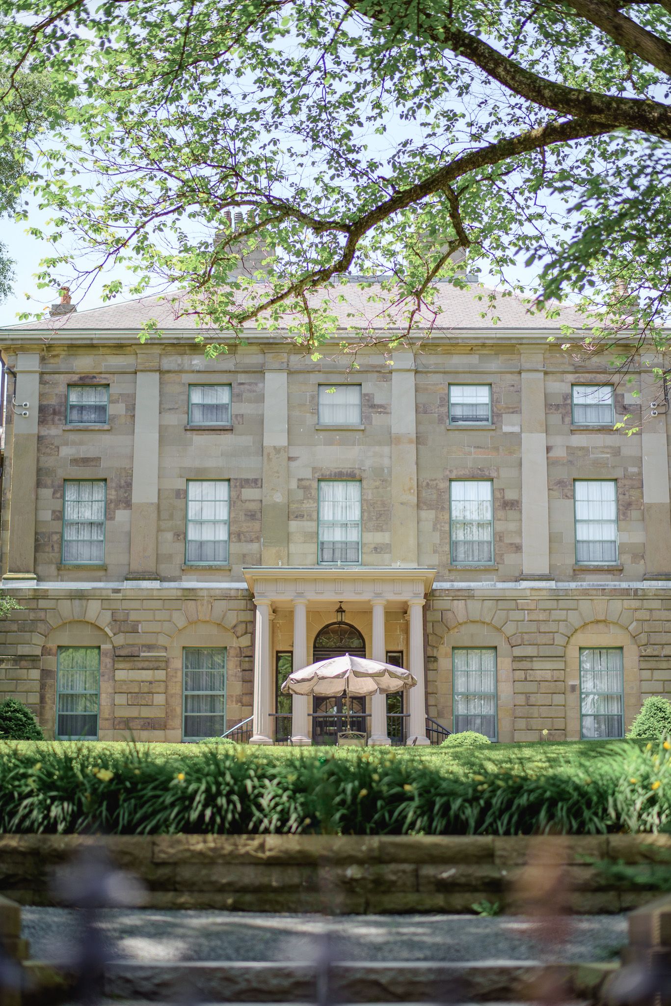 Historic stone building with columned entrance framed by lush green tree branches in summer.