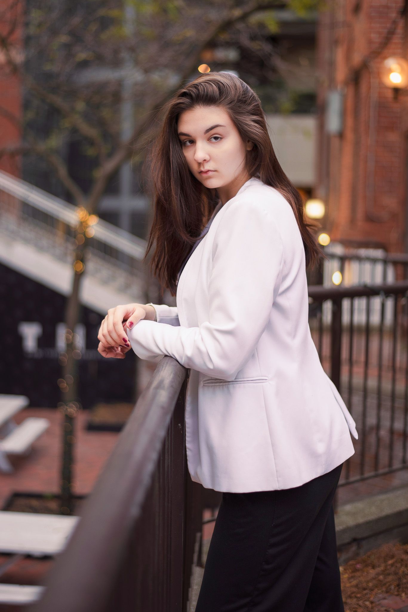 Young brunette woman in white blazer leaning on railing on urban outdoor balcony.