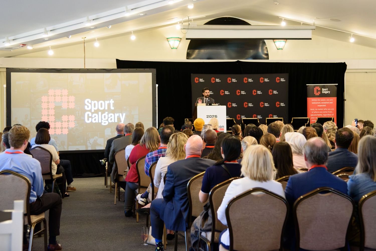 Sport Calgary conference presentation with speaker at podium addressing a large seated audience in a bright event hall.