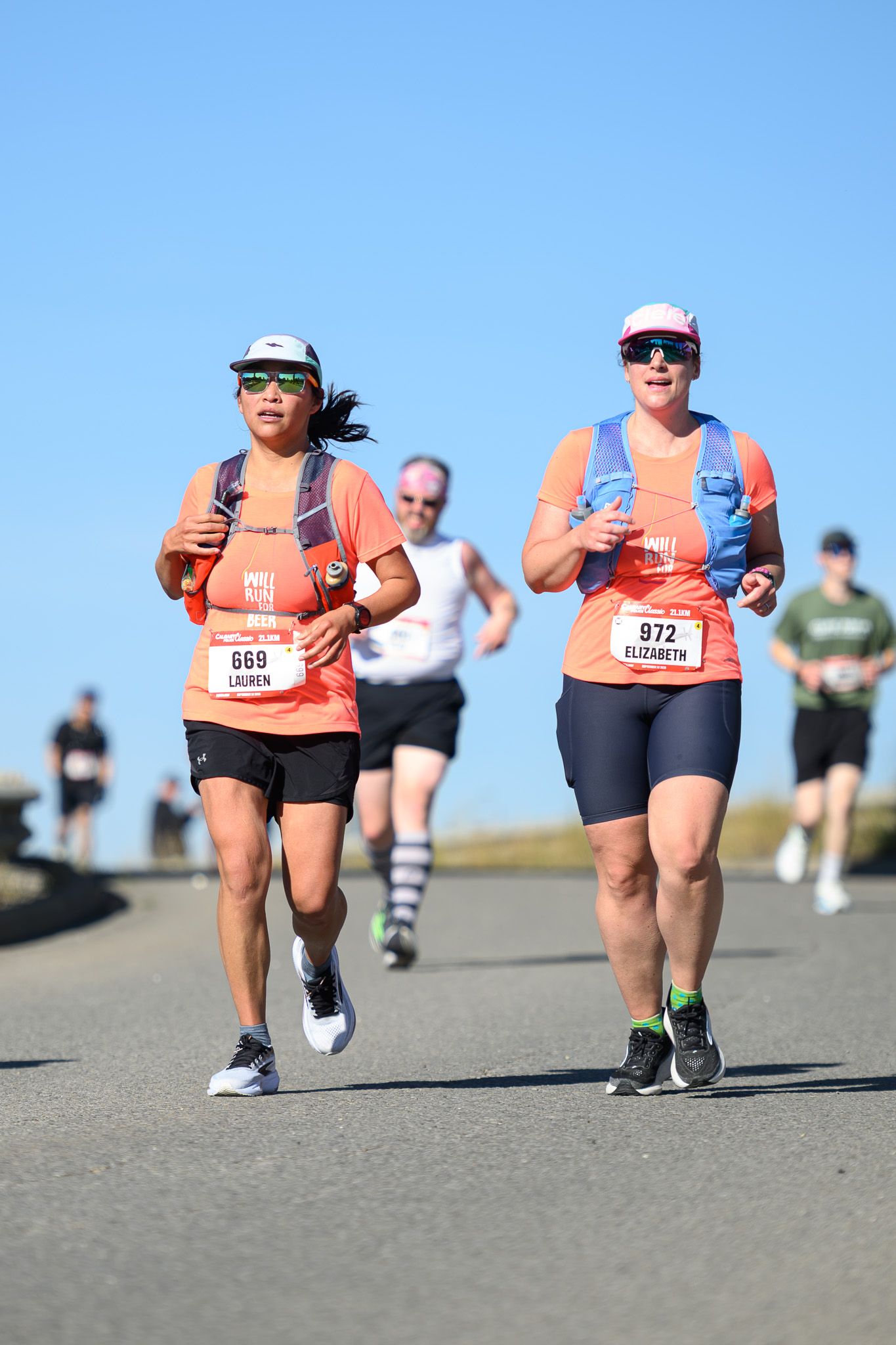Two women, Lauren #669 and Elizabeth #972, running in a road race wearing matching coral shirts on a sunny day.