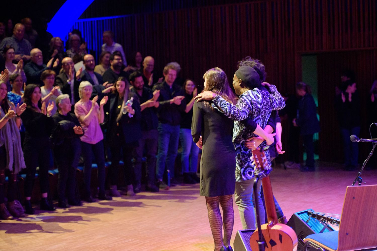 Two performers share an embrace on stage during a live music concert with audience applauding.