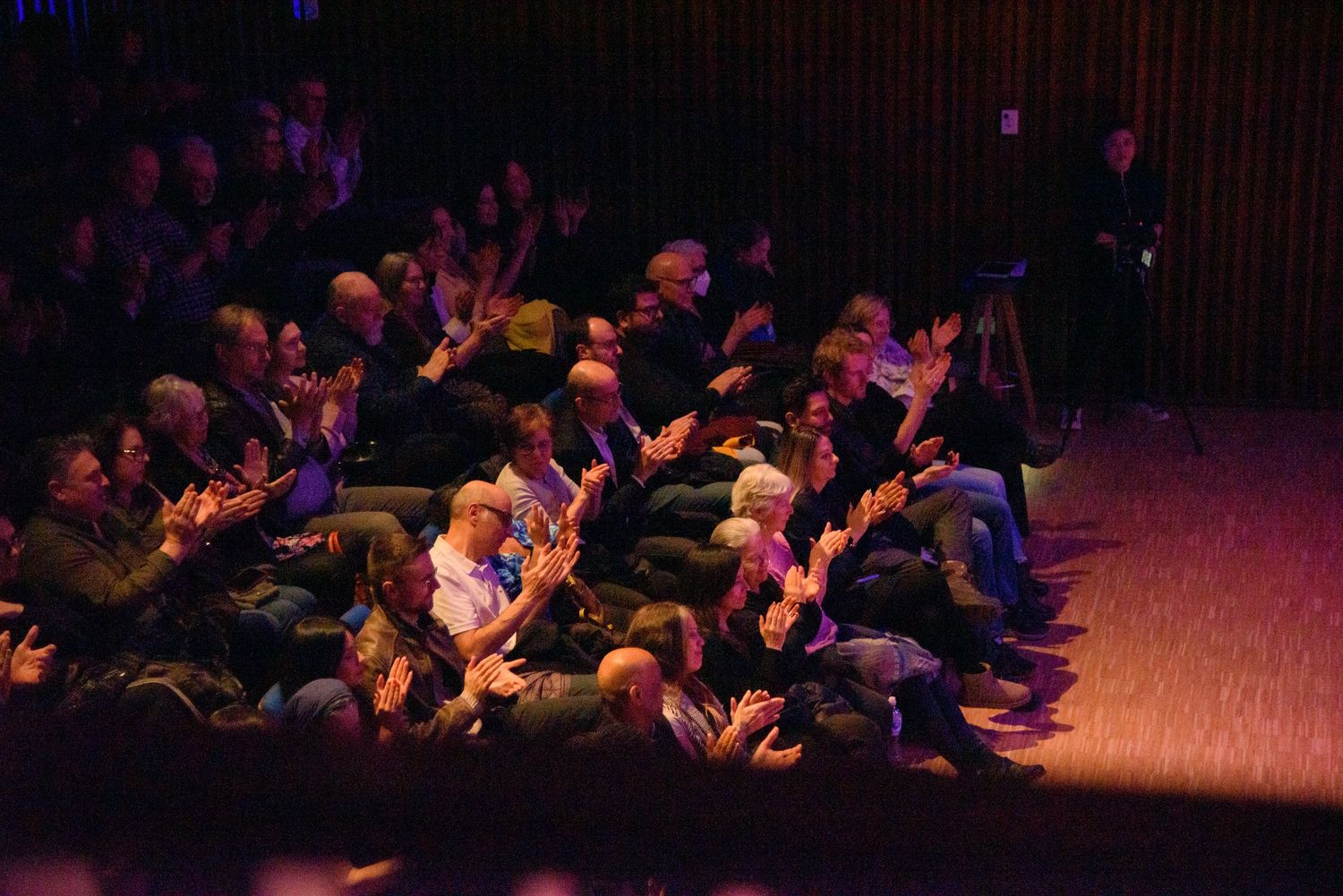Audience members clapping and applauding in a dimly lit theater during a live performance event.