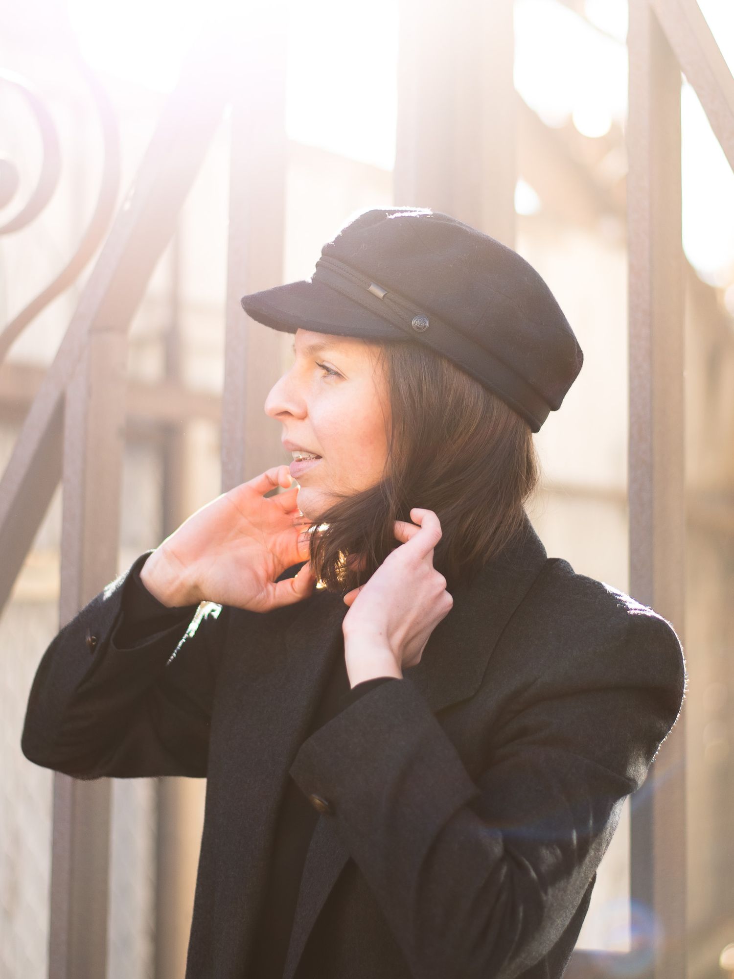 Brunette woman in black coat and newsboy cap adjusting collar outdoors in bright sunlight.