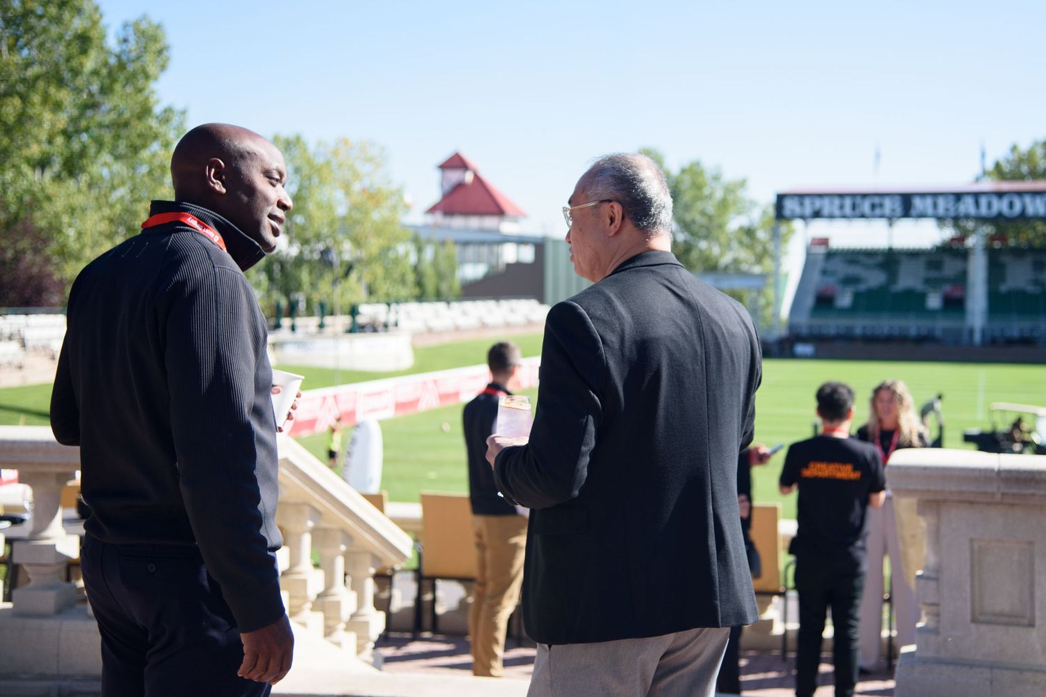 Two men conversing outdoors at Spruce Meadows equestrian venue on a sunny day.